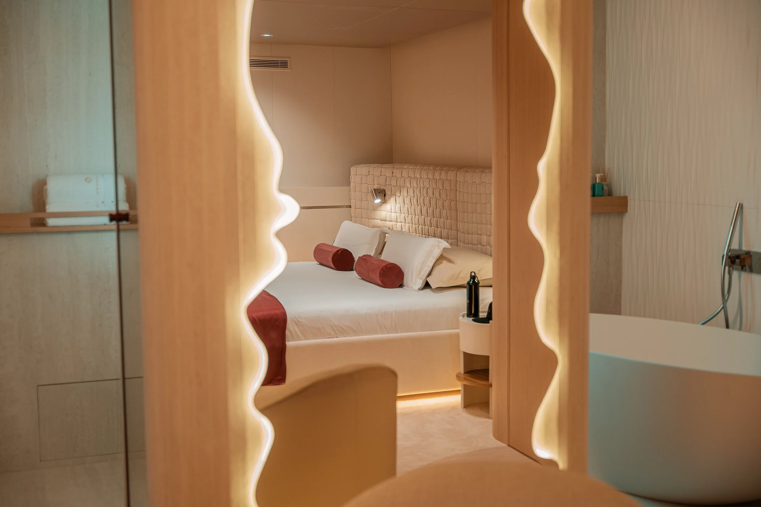 A cozy hotel bedroom with a large bed, beige tufted headboard, white and maroon pillows, and bedside lamps, viewed through a decorative mirror with wavy edges.