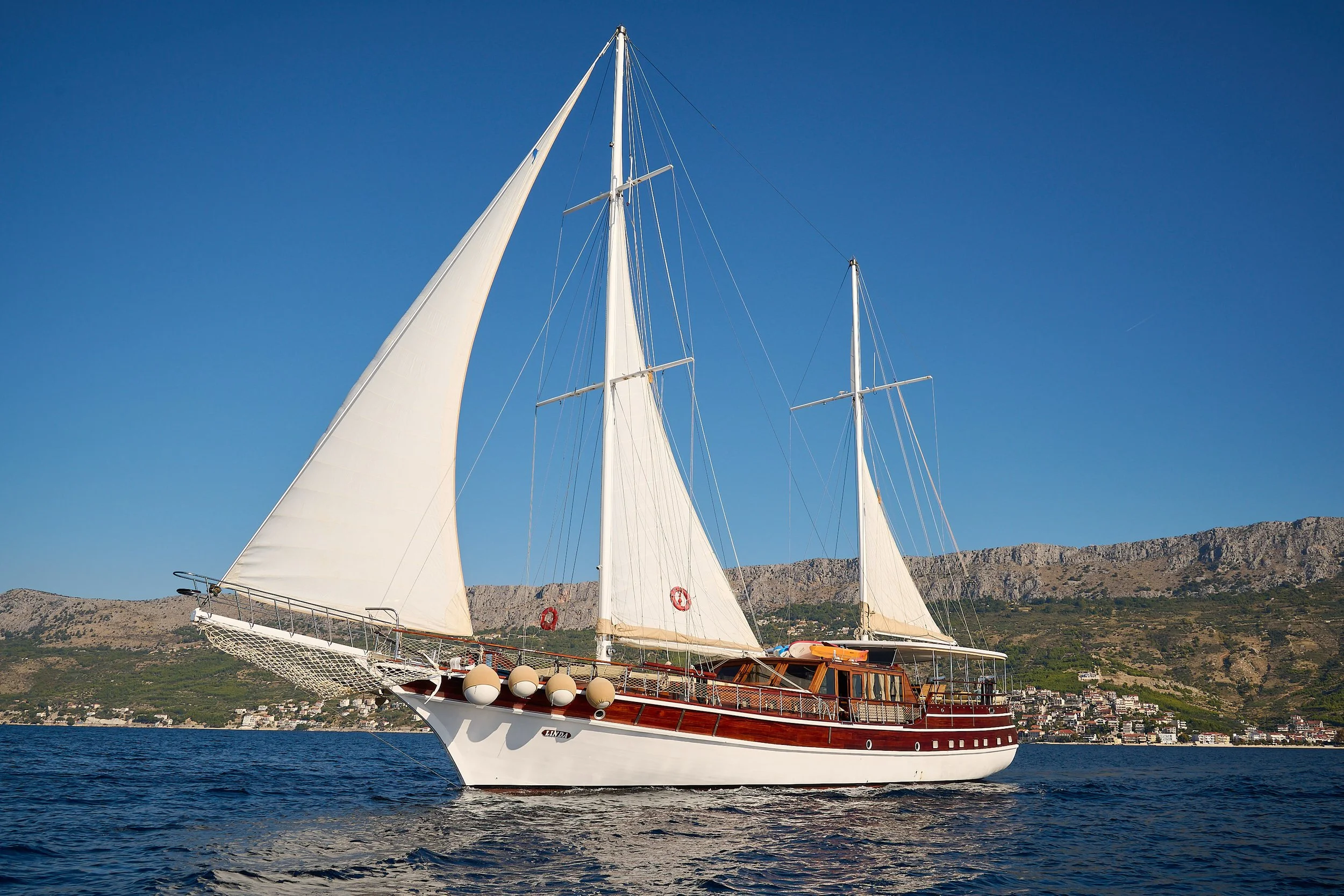 A sailing yacht with white sails and a wooden hull navigating on the water with coastal hills and buildings in the background under a clear blue sky.
