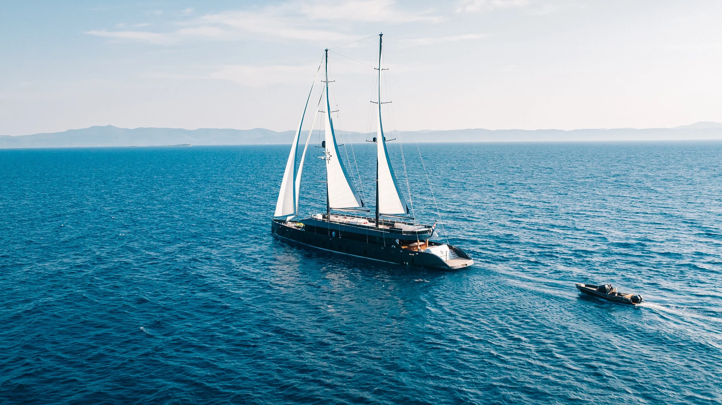 A large black sailboat with white sails on the open water, accompanied by a small motorboat to its side, with distant mountains on the horizon.