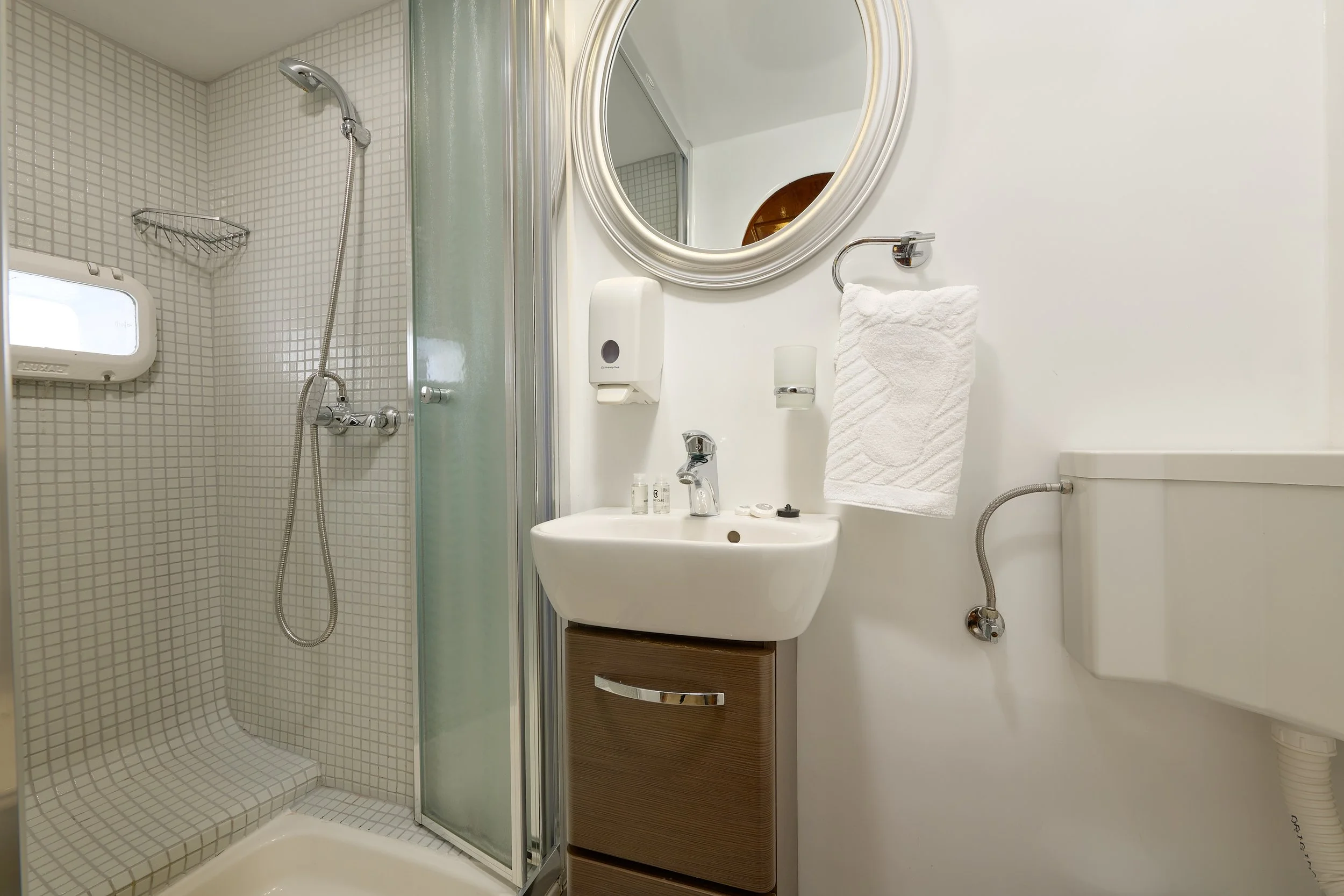 Small bathroom with a corner shower stall, a white oval mirror, a wall-mounted sink with a wooden cabinet underneath, and a white toilet. Towel and soap dispensers are on the wall, and there is a small window in the shower area.