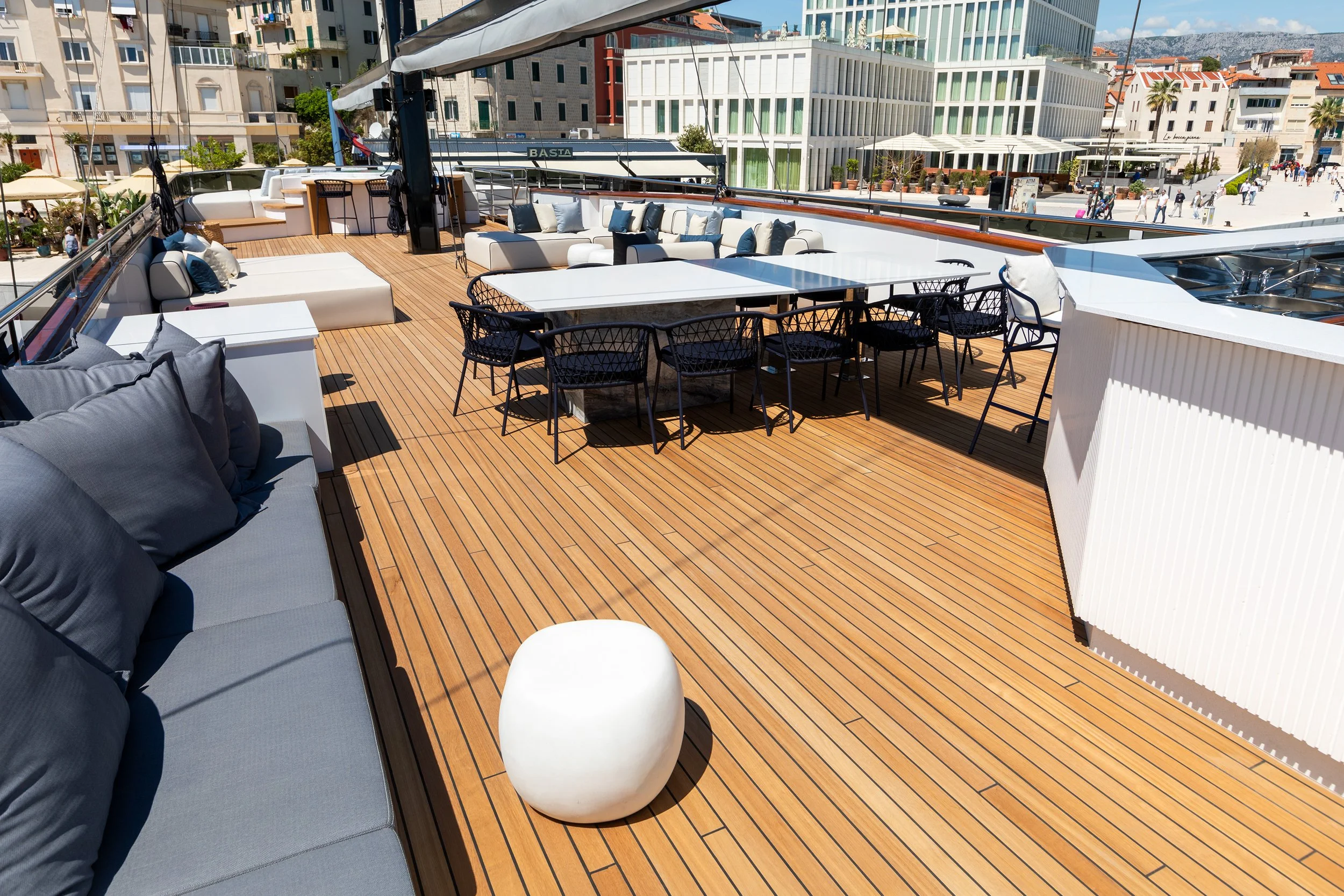 A yacht deck with outdoor furniture, including cushioned loungers, a white marble dining table with black chairs, and a white ceramic stool. There are buildings and people in the background, indicating a city waterfront setting.