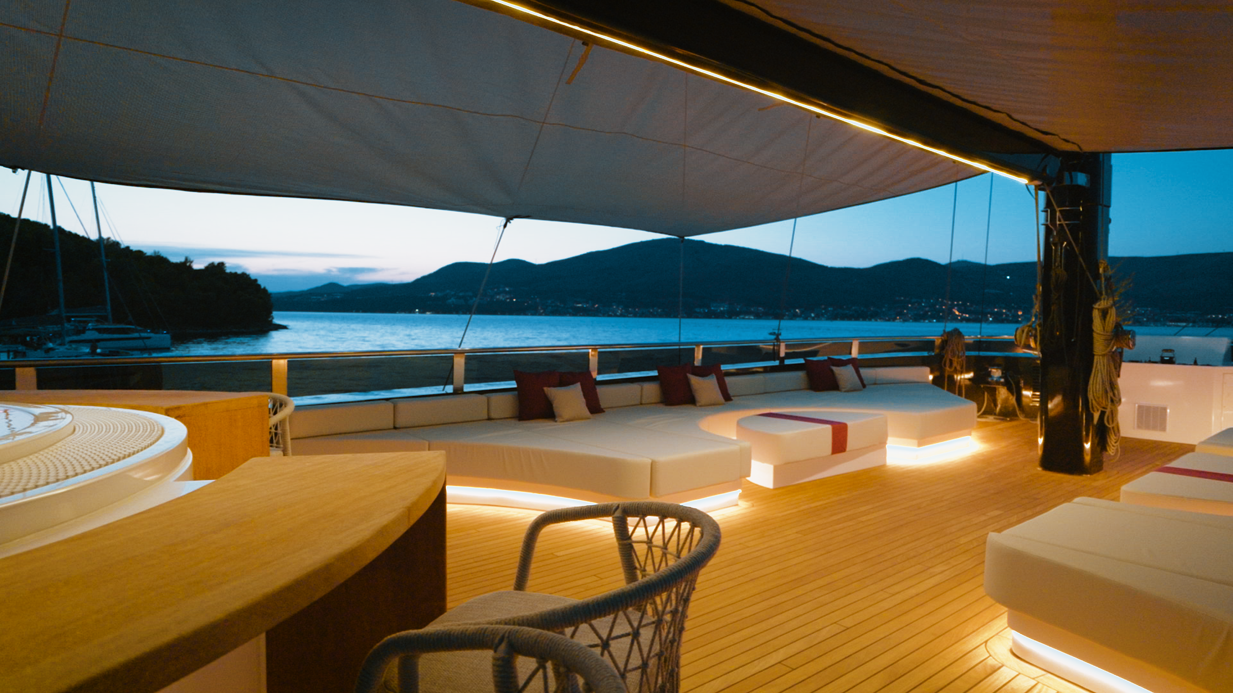 An outdoor lounge area on a yacht at dusk with white couches and pillows, a wooden deck, and a view of water and mountains in the background.