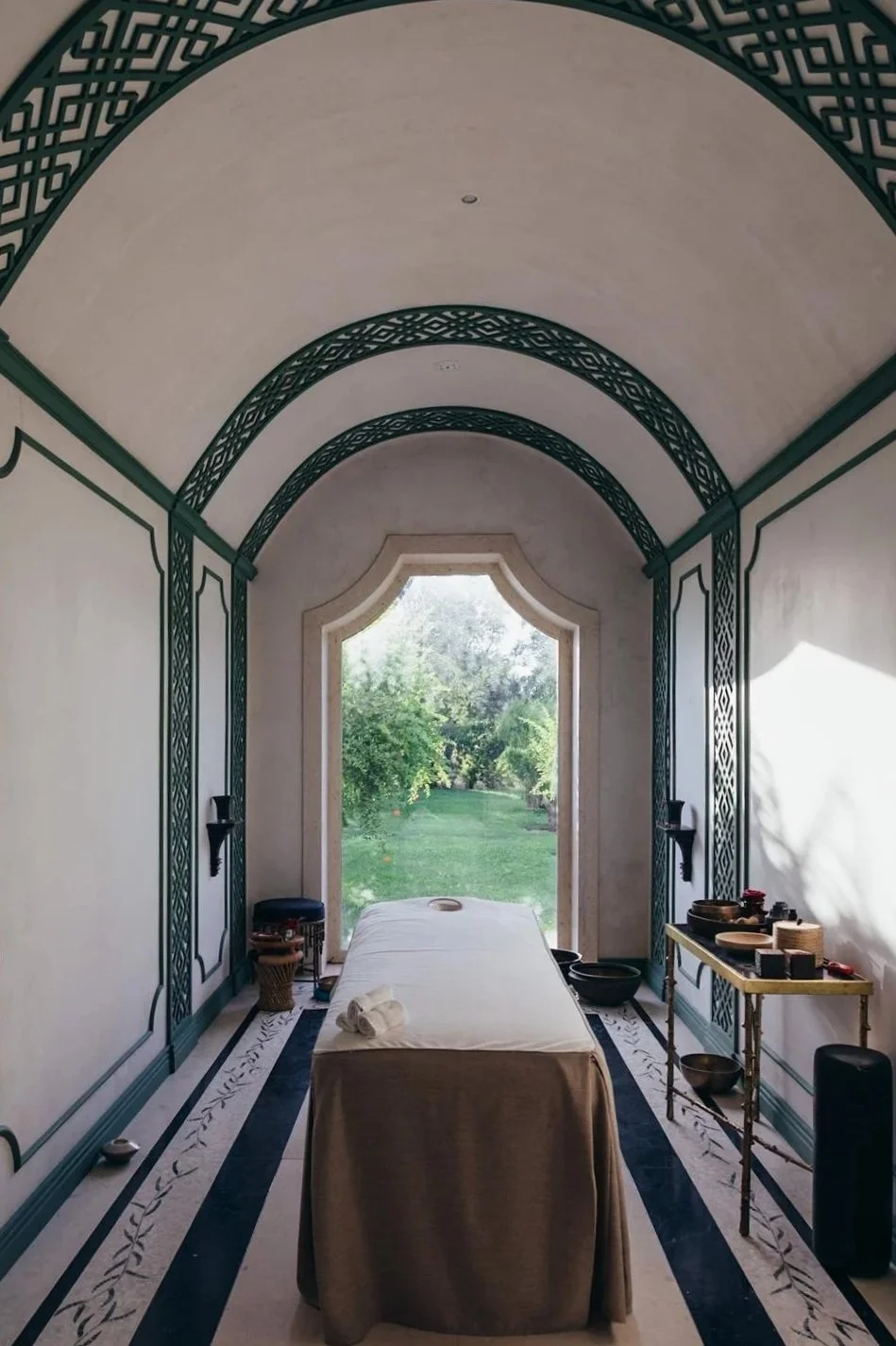 A massage room with a massage table covered in a beige sheet, positioned facing a large arched window overlooking a lush green garden. The room has decorative teal and white patterned archways along the ceiling and walls, with black wall-mounted candle holders. To the right, a small gold table holds massage tools and bowls, and a black speaker is on the floor. There is a small stool and a basket with rolled towels near the head of the table.