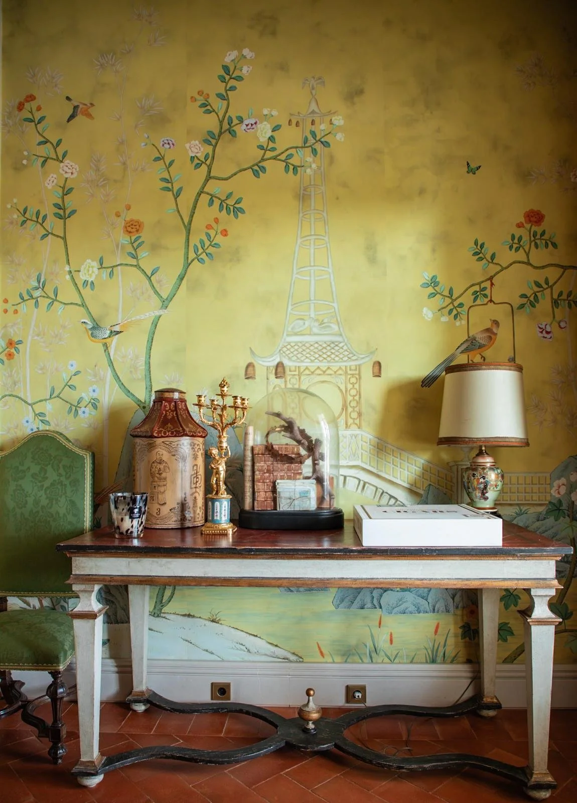Interior of a room with a decorative wall mural featuring birds, branches, flowers, and a tower. A wooden table holds a lamp, a glass display case, and decorative items including a candlestick and a small figurine. A green chair is partially visible on the left, and a lit table lamp is on the right.