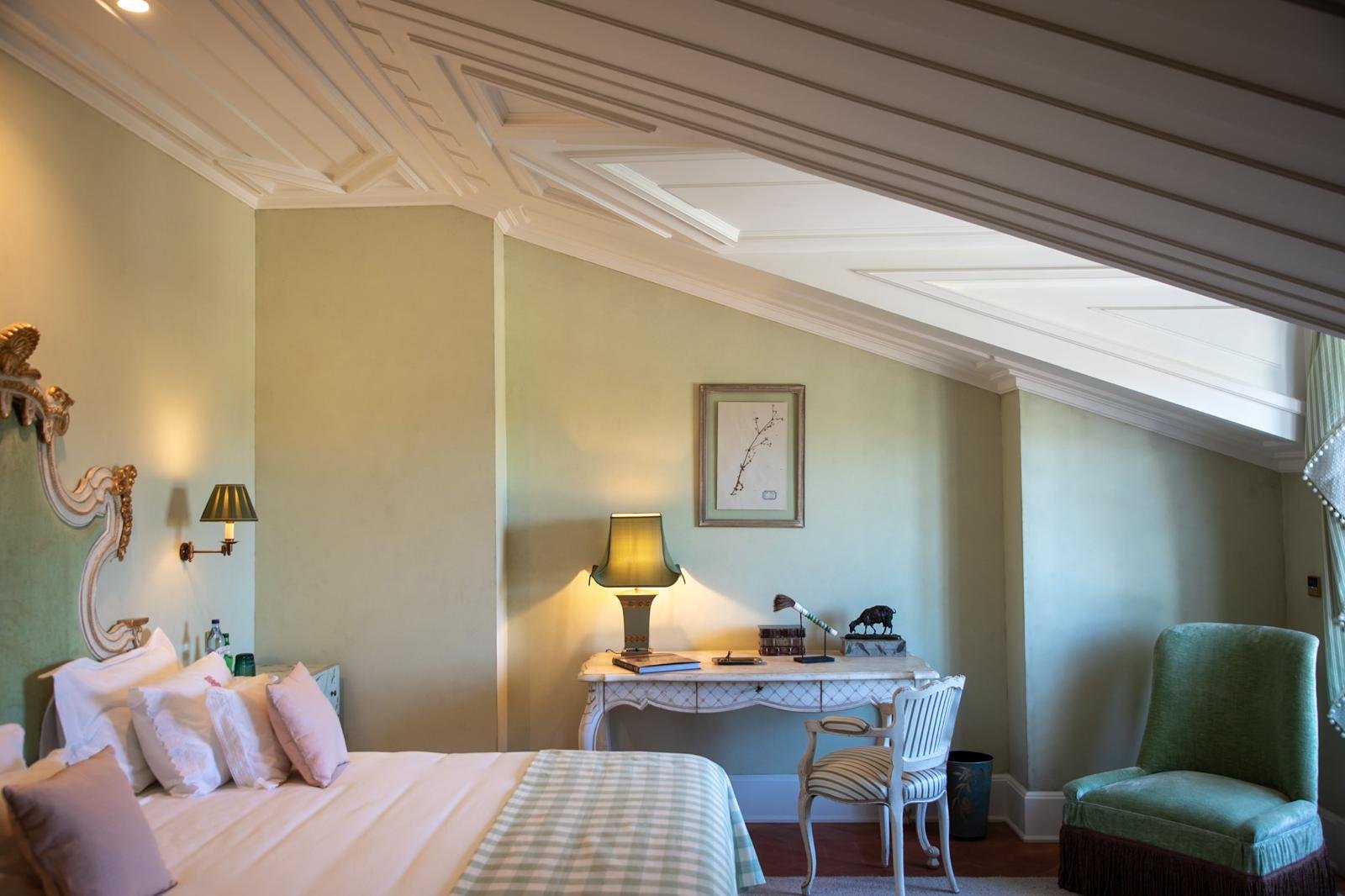 A cozy hotel room with a bed featuring decorative pillows and a distinctive ornate headboard. The room has soft green walls, a desk with a lamp and decorative items, and a comfortable green armchair. Natural light filters in through a skylight and window with curtains.