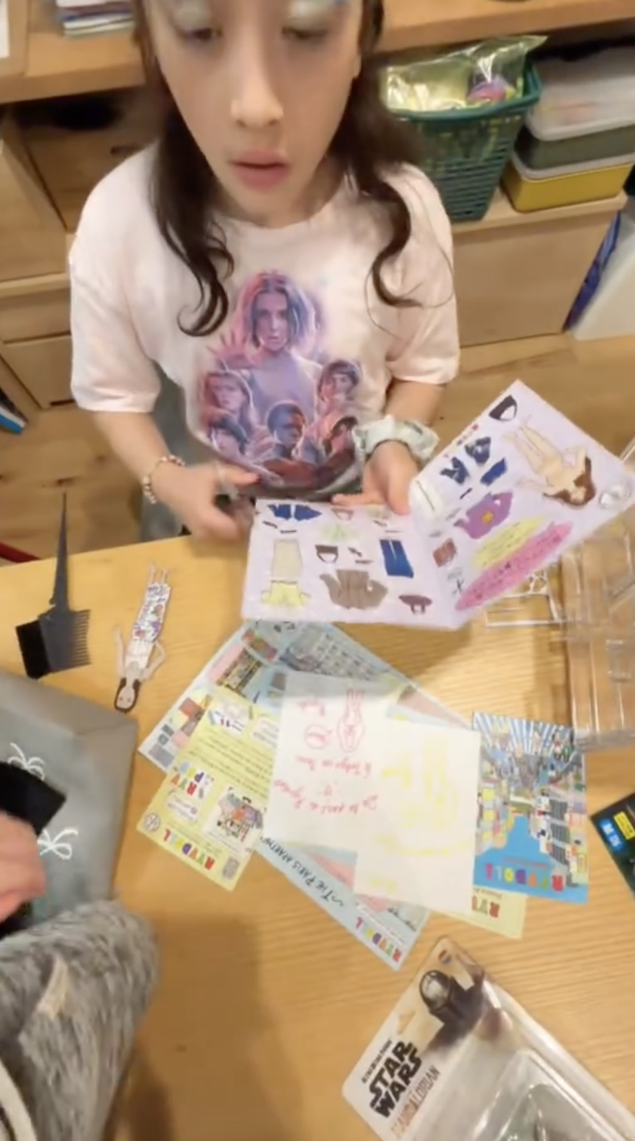 A young woman is looking at a colorful brochure or magazine on a wooden table, surrounded by various papers, flyers, and a comb. She is wearing a light-colored T-shirt featuring characters from a popular TV show.