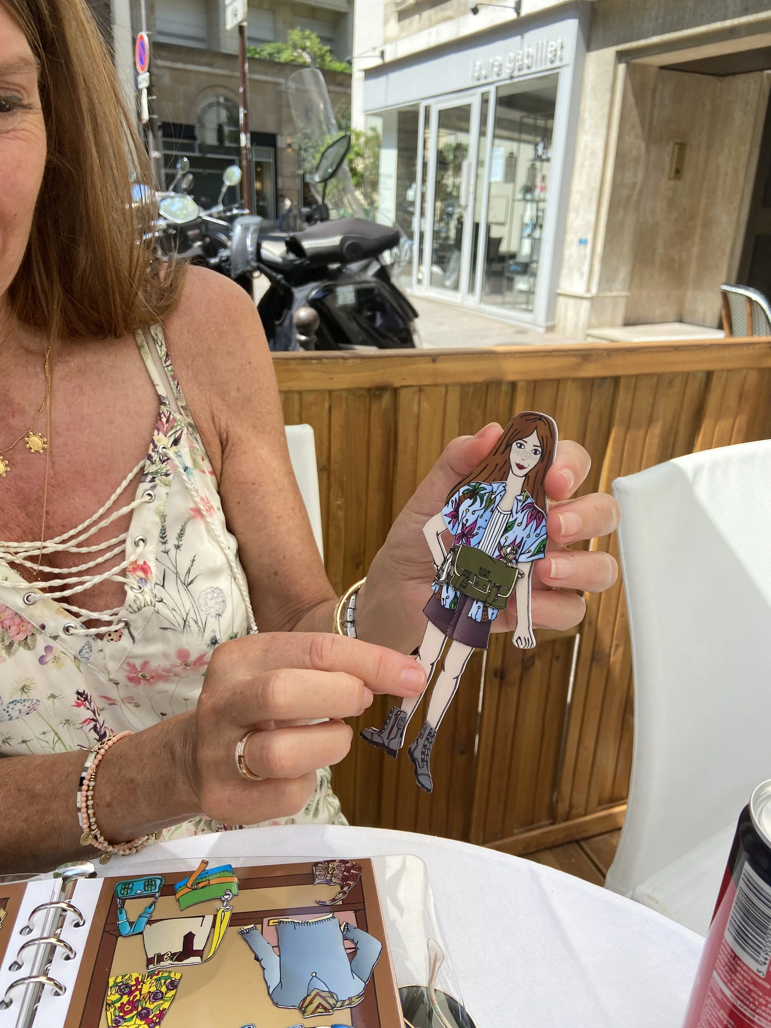 A woman holding a paper doll illustration of a girl with long brown hair, wearing a patterned shirt, purple skirt, and combat boots at an outdoor restaurant table.