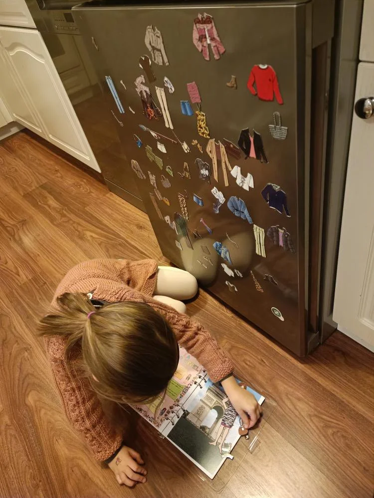 A young girl lying on the wooden floor, looking at an open book, next to a fridge covered with clothing magnets forming a collage.