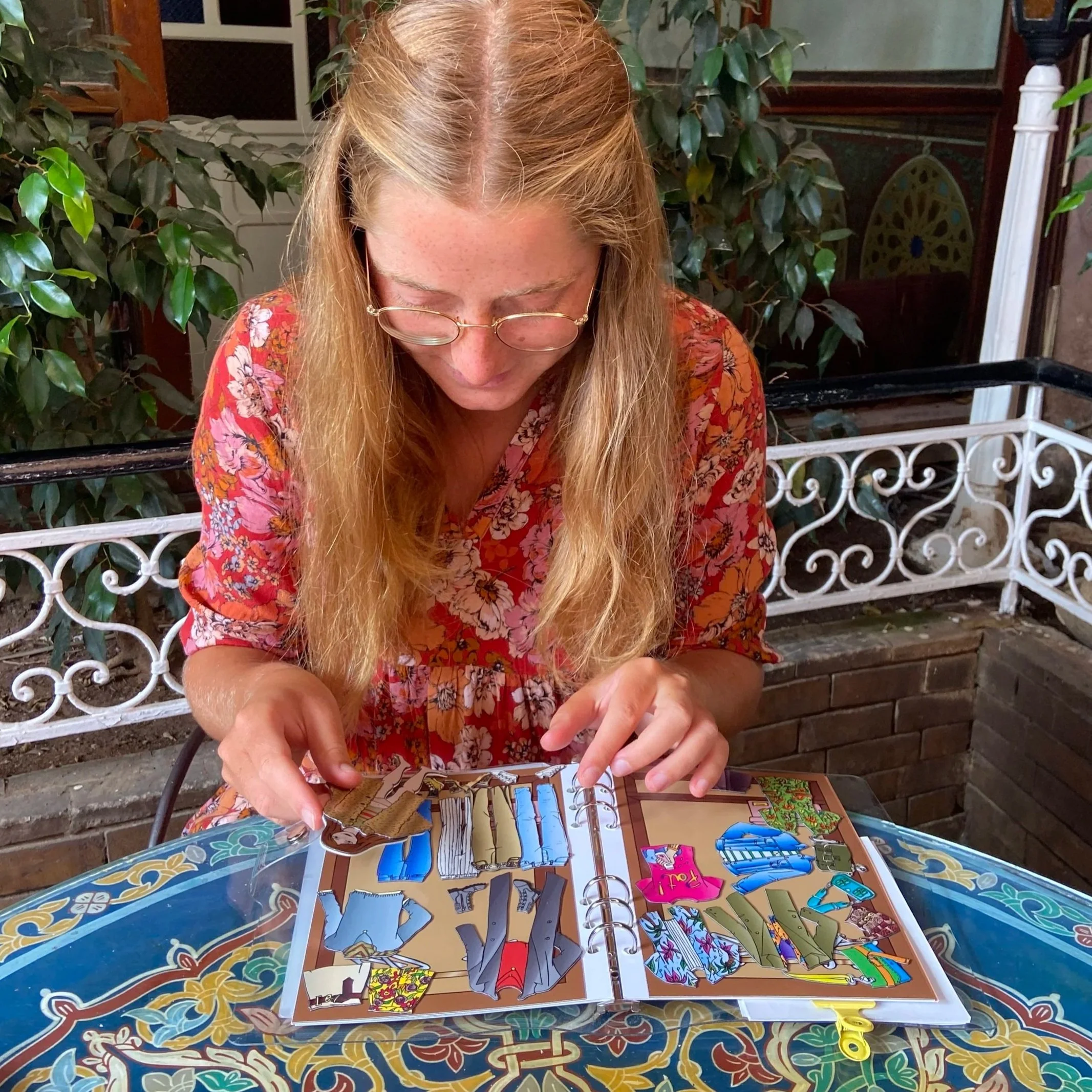 A woman with long blonde hair and glasses, wearing a floral orange top, sits at a table looking at a colorful book or magazine with various illustrations. She appears to be pointing or turning pages. The table has a decorative multicolored surface, a