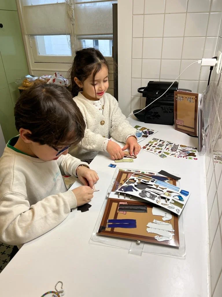 Two children are working on a paper craft project at a white counter in a kitchen. They are cutting and assembling paper pieces with various pictures and colors. A window, some snacks, and a printer are visible in the background.