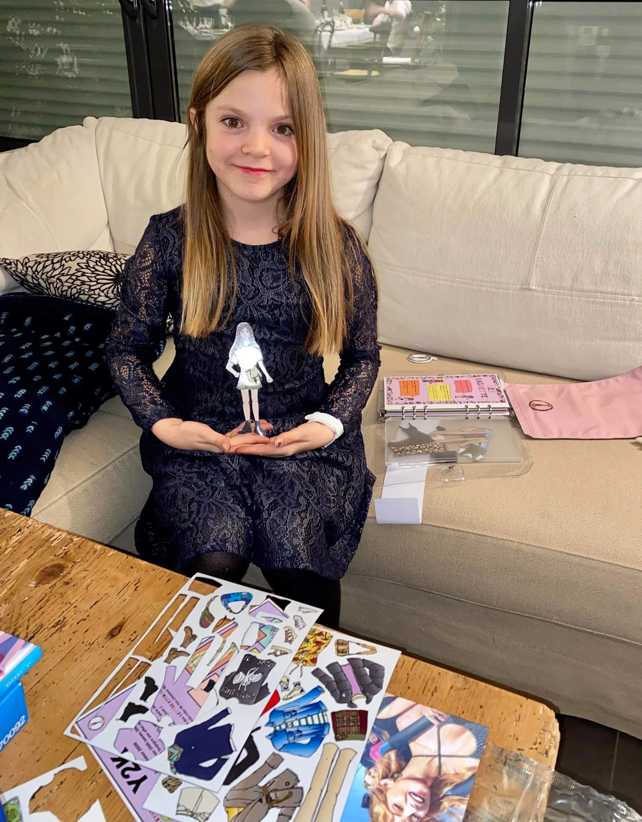 A young girl with long brown hair, wearing a dark lace dress, sitting on a beige couch in a living room. She is holding an animated light-up figurine of a girl. In front of her, on a wooden table, there are colorful paper cutouts and craft supplies. 