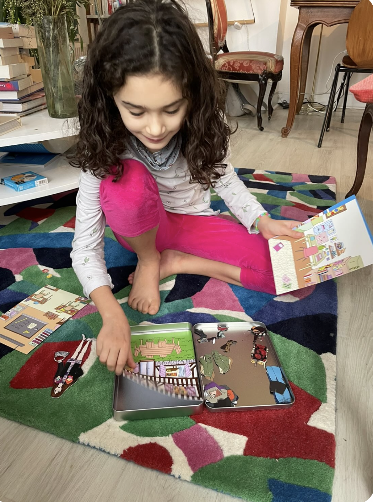 A young girl sitting cross-legged on a colorful patterned rug, playing with wooden doll puzzle pieces and a children's book open.