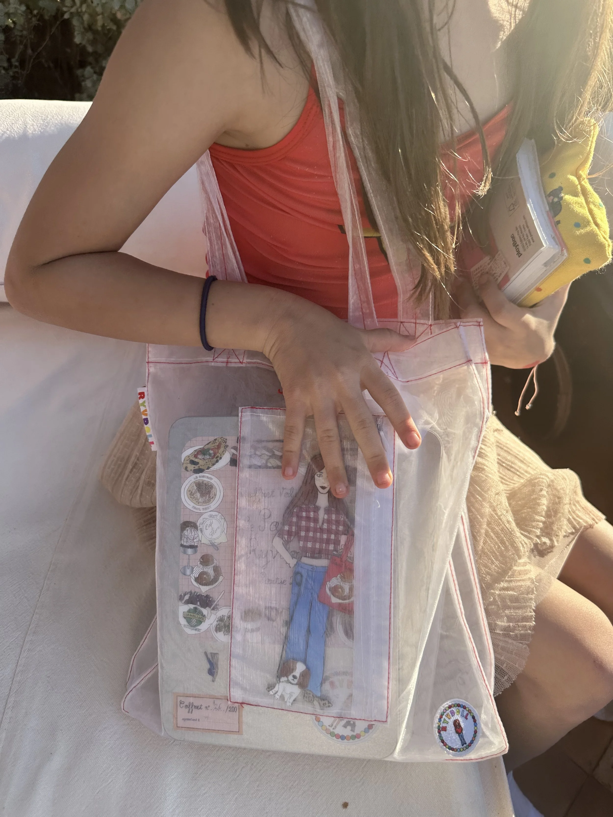 Person sitting at a table, wearing a red top, with a handmade fabric art project of a girl in a red checked shirt, blue jeans, and dog on the table in front of her. The person is holding a book and a yellow cloth, and the table has a white cover.