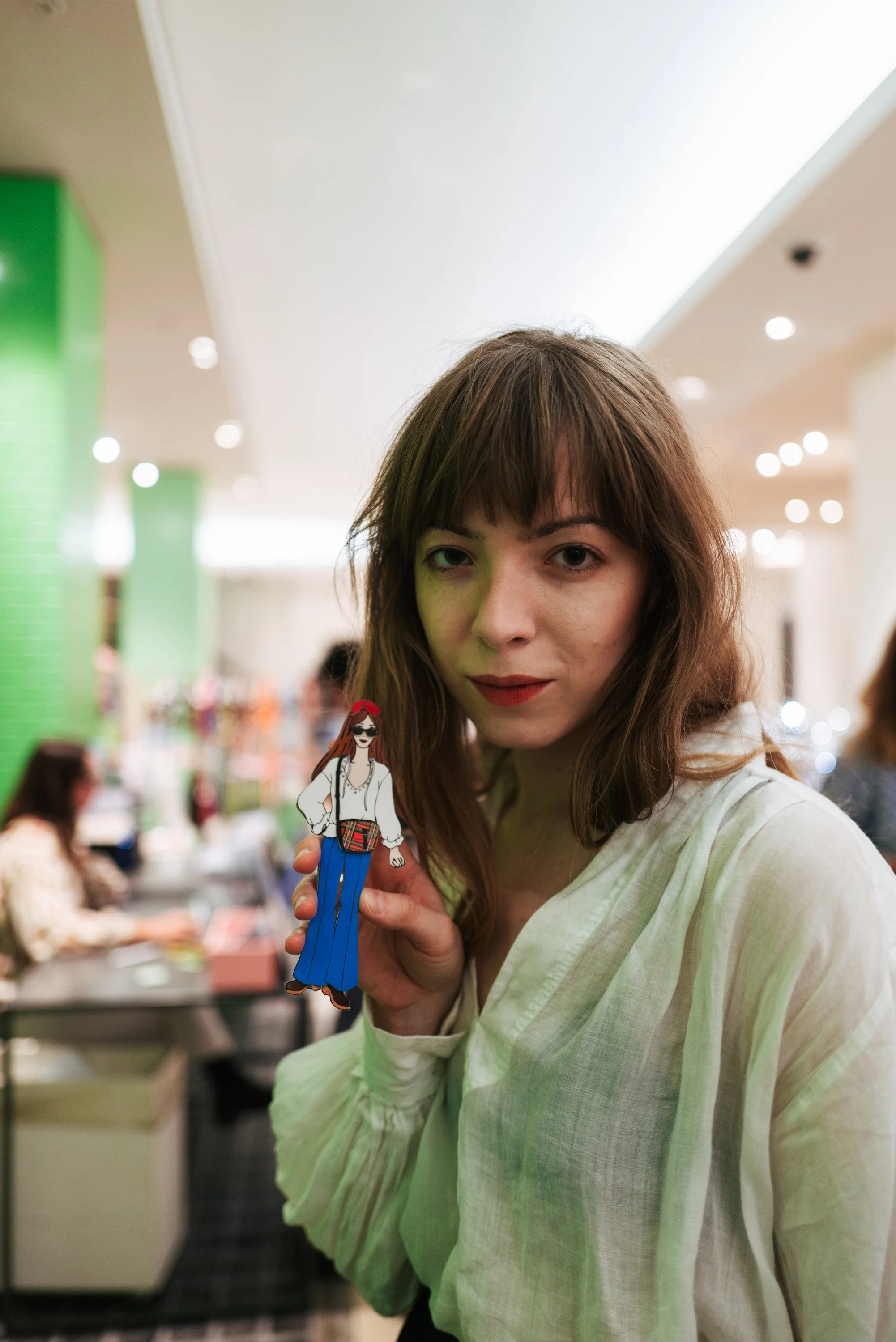 A young woman with brown hair and a white shirt holding a cartoon illustration of a woman with long hair, glasses, and colorful clothing in a busy indoor setting.