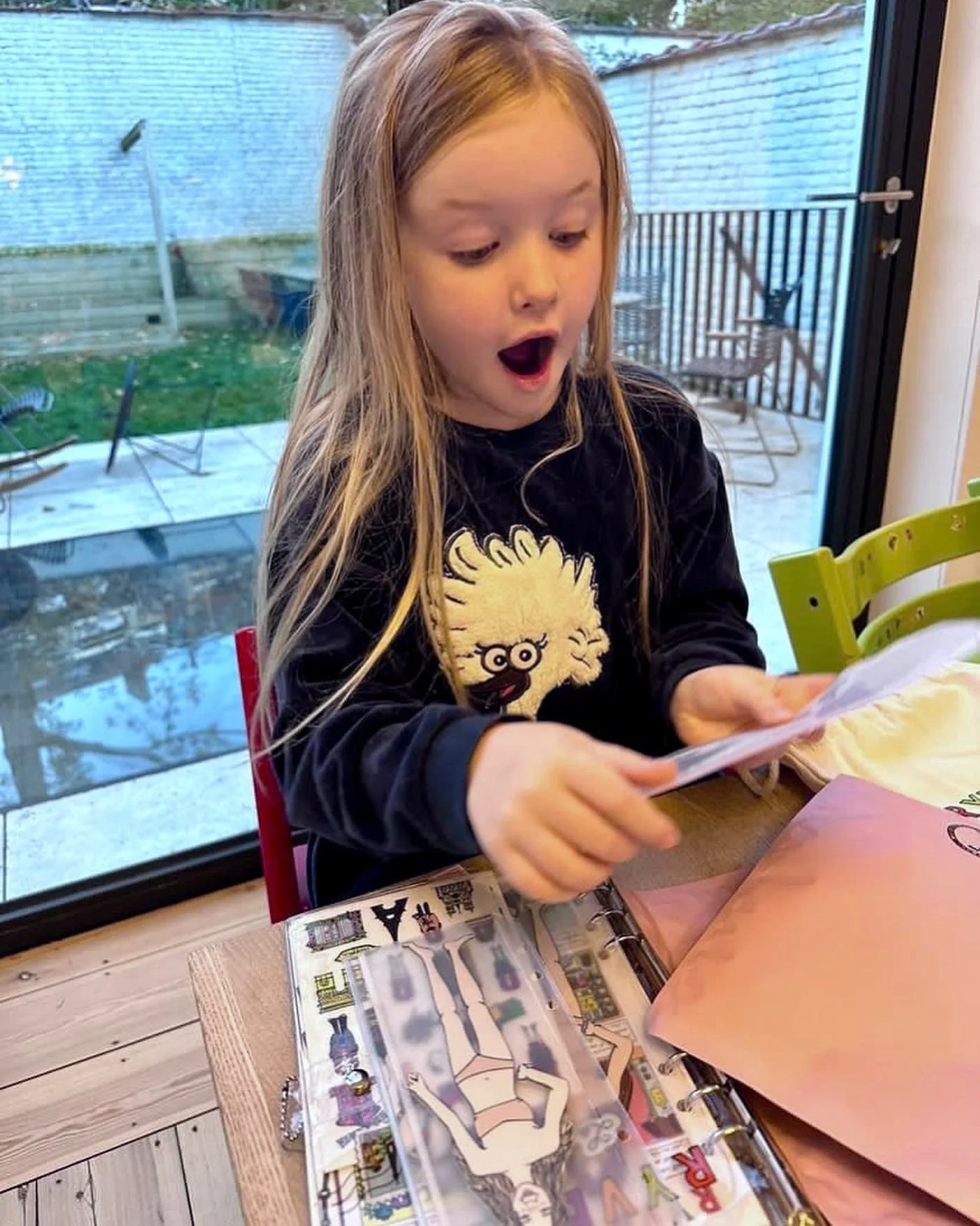 A young girl with long blonde hair sitting at a table, looking at a piece of paper with an expression of surprise while opening a plastic photo album with a picture of a woman in a bikini.