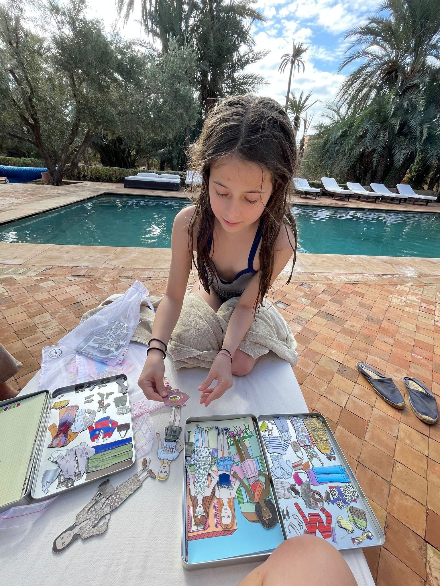 A young girl sitting by a pool, organizing paper dolls and cutouts of clothing on a table outdoors, with pool chairs and trees in the background.