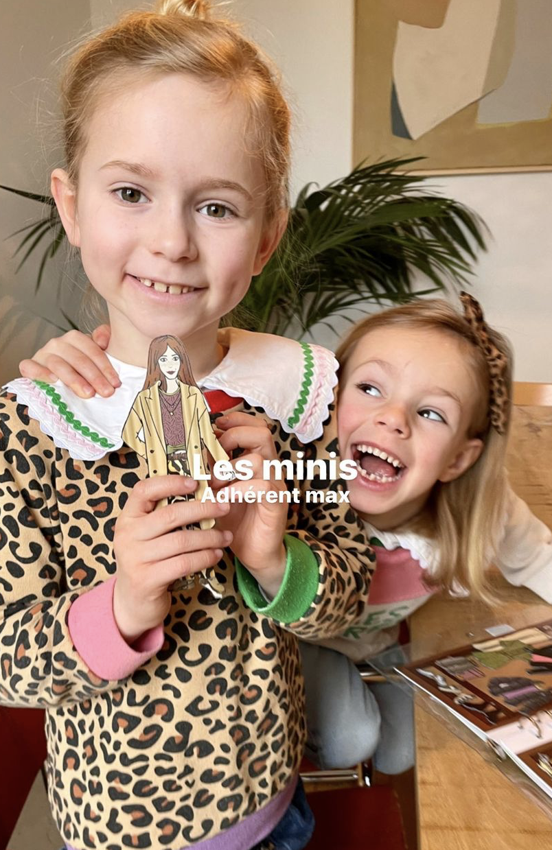 Two young girls smiling and holding a paper doll in a room with a plant and artwork in the background. One girl is wearing a leopard print hoodie and the other is wearing a white shirt with a leopard print headband.