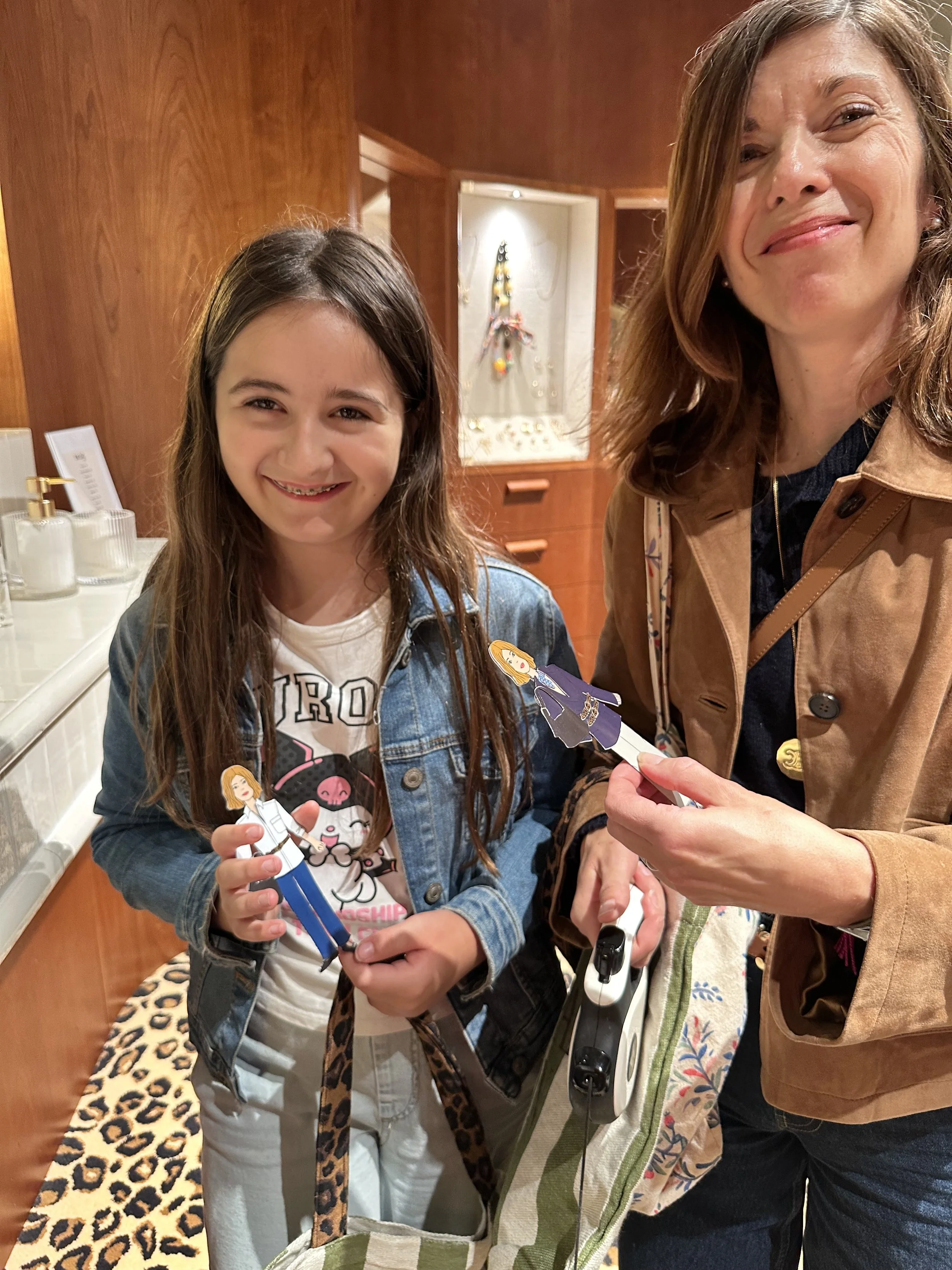 A smiling young girl and an older woman standing inside a warmly lit room, both holding paper cutouts of fictional characters. The girl has long brown hair, wears a denim jacket and a white t-shirt, and has a leopard-print bag strap across her chest.