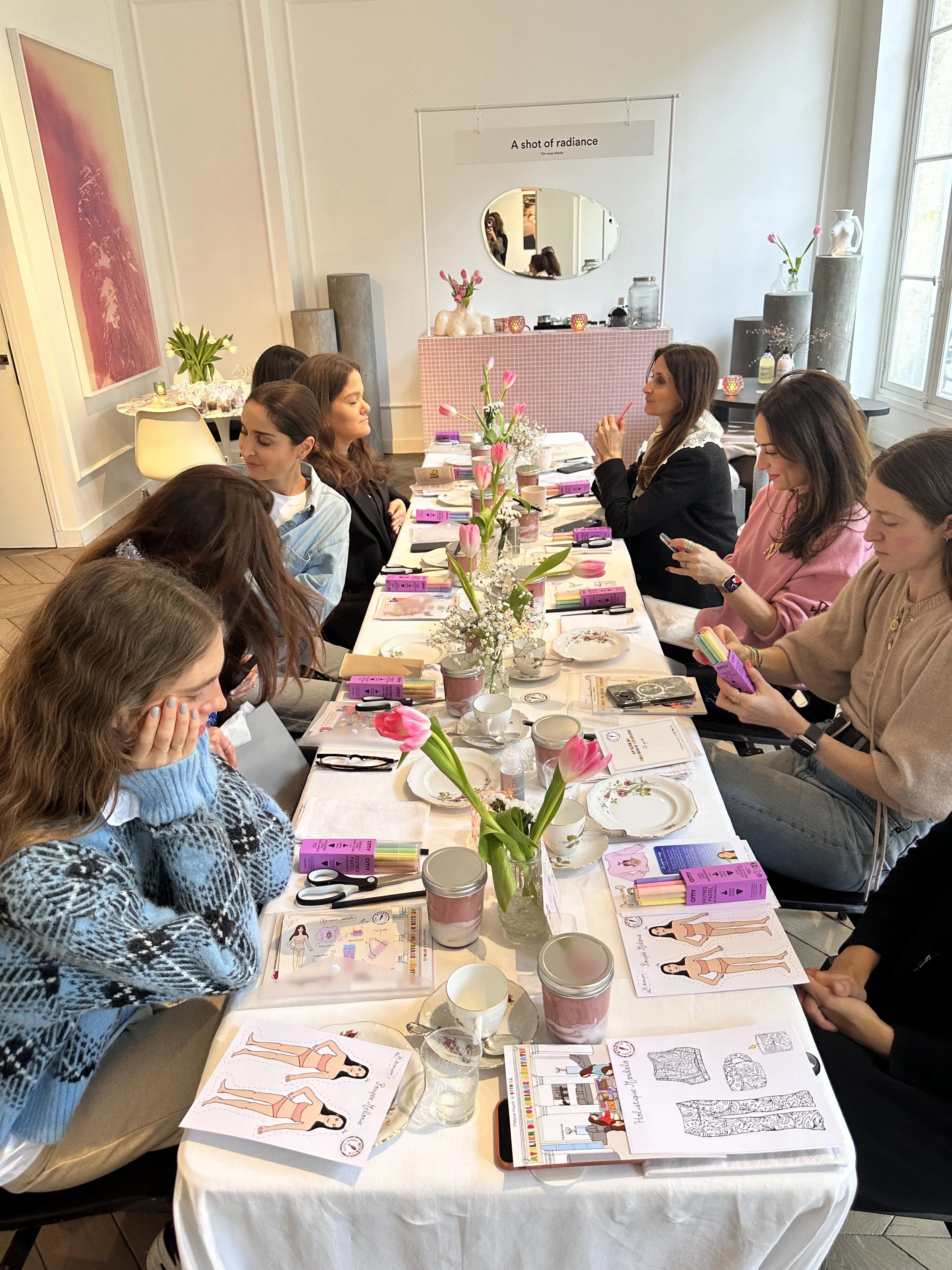 Women gathered around a long table for a crafting workshop, with pink tulips and various art supplies, in a brightly lit room with large windows.