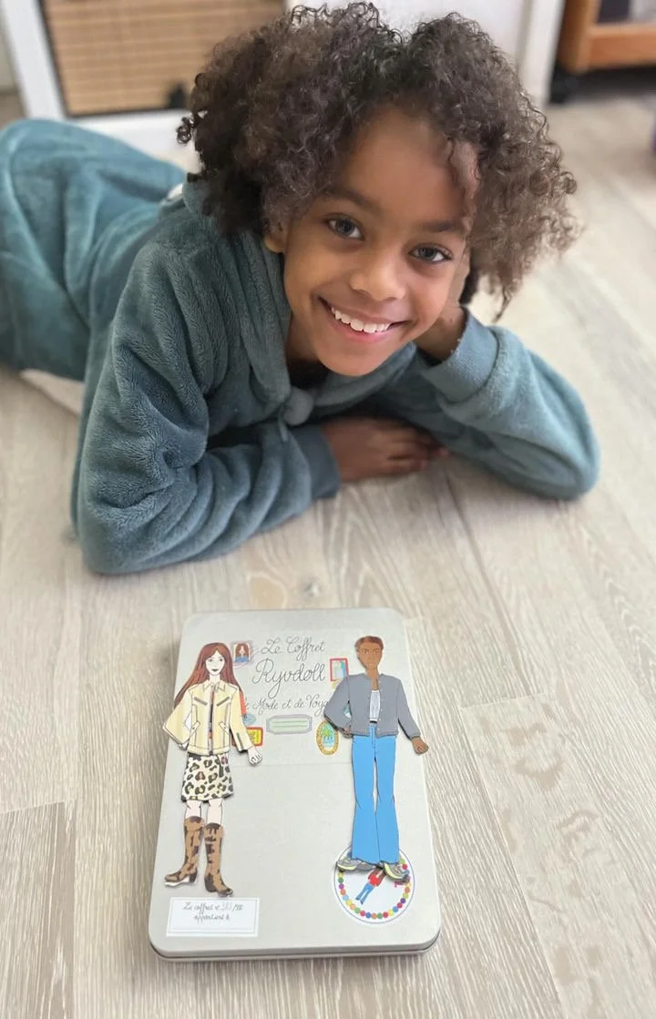 A young girl lying on a wooden floor, smiling at the camera, with illustrated paper dolls and a white box in front of her.