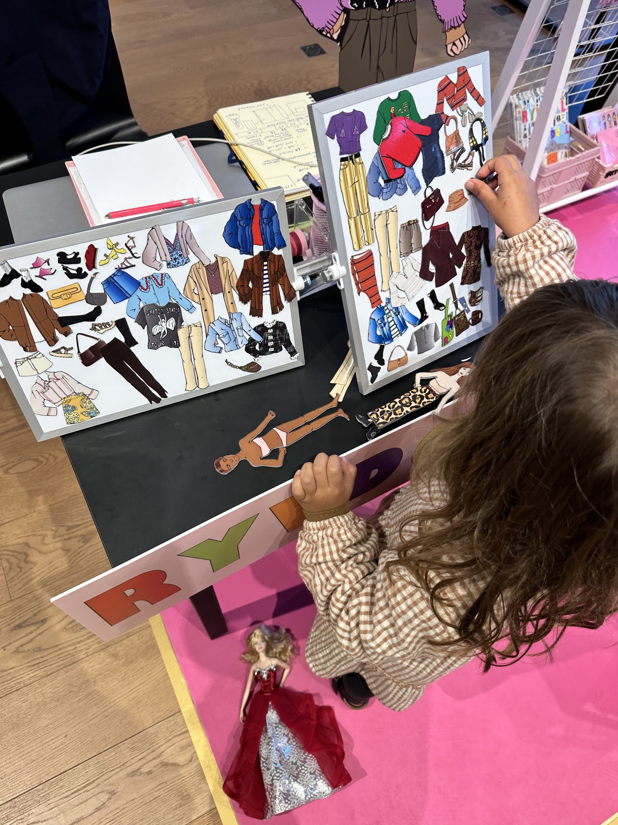 A young girl is looking at two display boards with cutout fashion clothing and accessories, while a Barbie doll in a red gown is on the pink floor nearby.