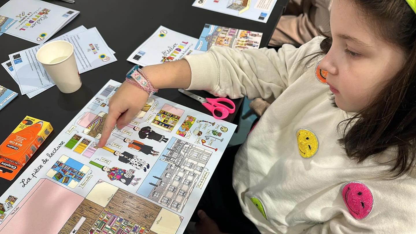 A young girl with brown hair, wearing a cream sweatshirt with smiley face patches, points at an educational poster about a library and reading. The table has scissors, a box of juice, and various papers and flyers.