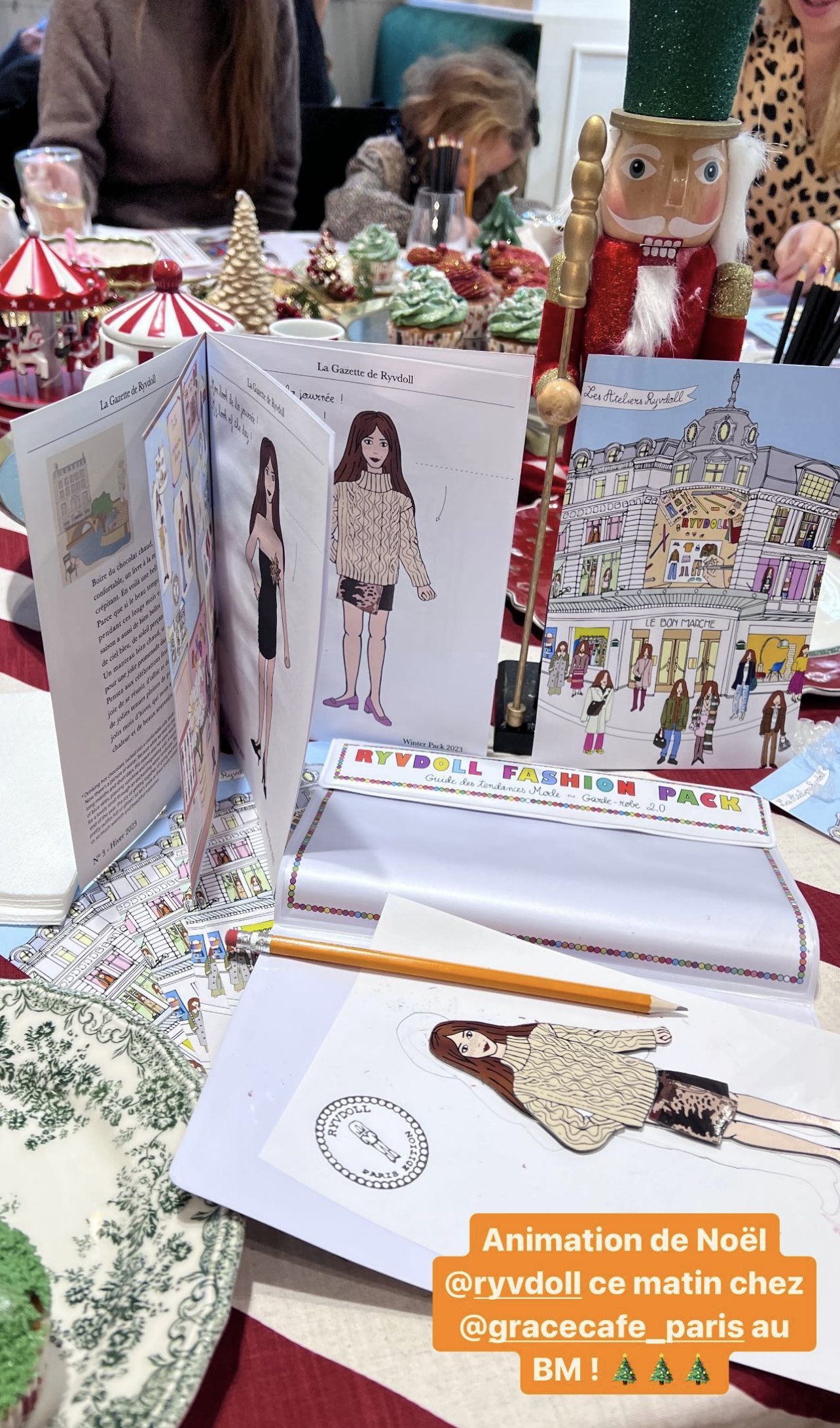 Christmas-themed table with RyvDoll fashion pack, illustrated fashion magazine, toy nutcracker figure, cupcakes with green frosting, and a Christmas village decoration, surrounded by people at a holiday event.