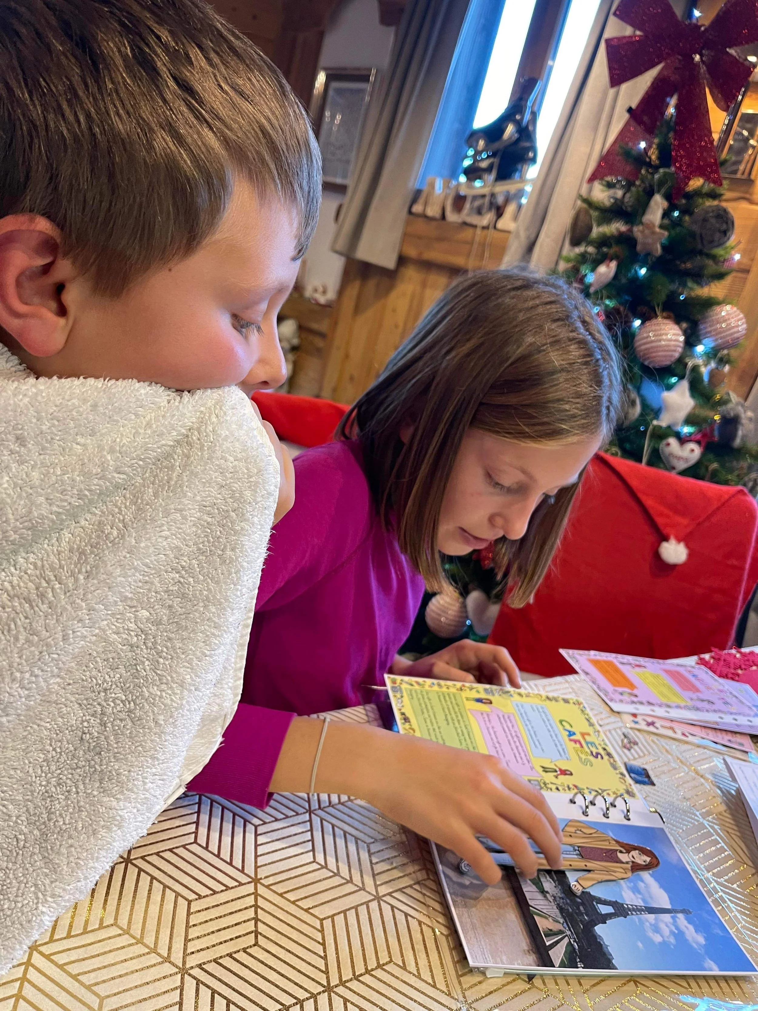 A boy and girl sitting at a table reading a scrapbook or photo album, with a decorated Christmas tree in the background.