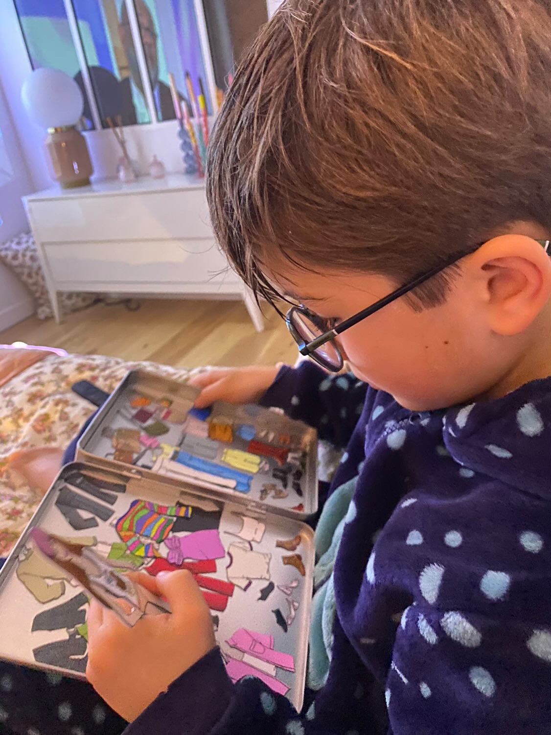 A young boy with glasses looking at a board game with cardboard cutouts of clothing and accessories inside a room with a dresser and decor.
