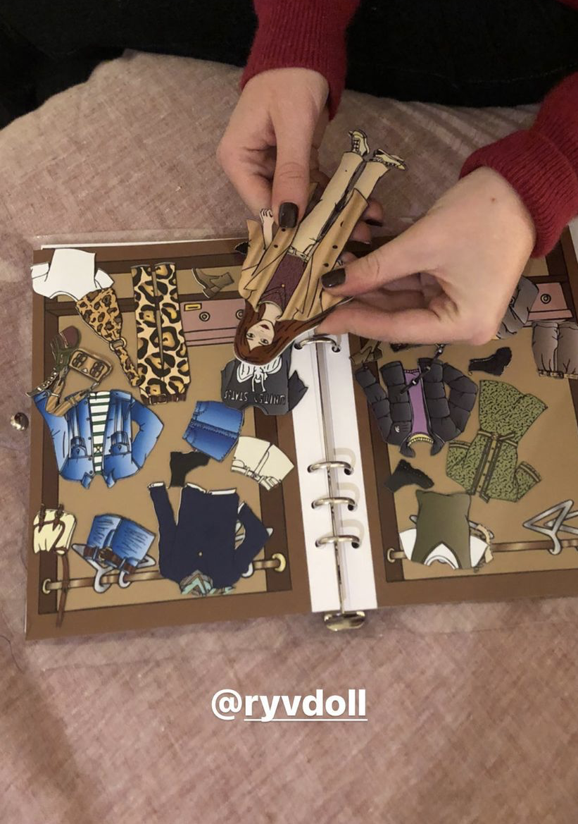 A person with black-painted nails and wearing a red sweater is assembling a paper doll set with different outfits on a pink surface. The paper doll set is in an open binder showing various fashion styles including leopard print, striped, denim, and g