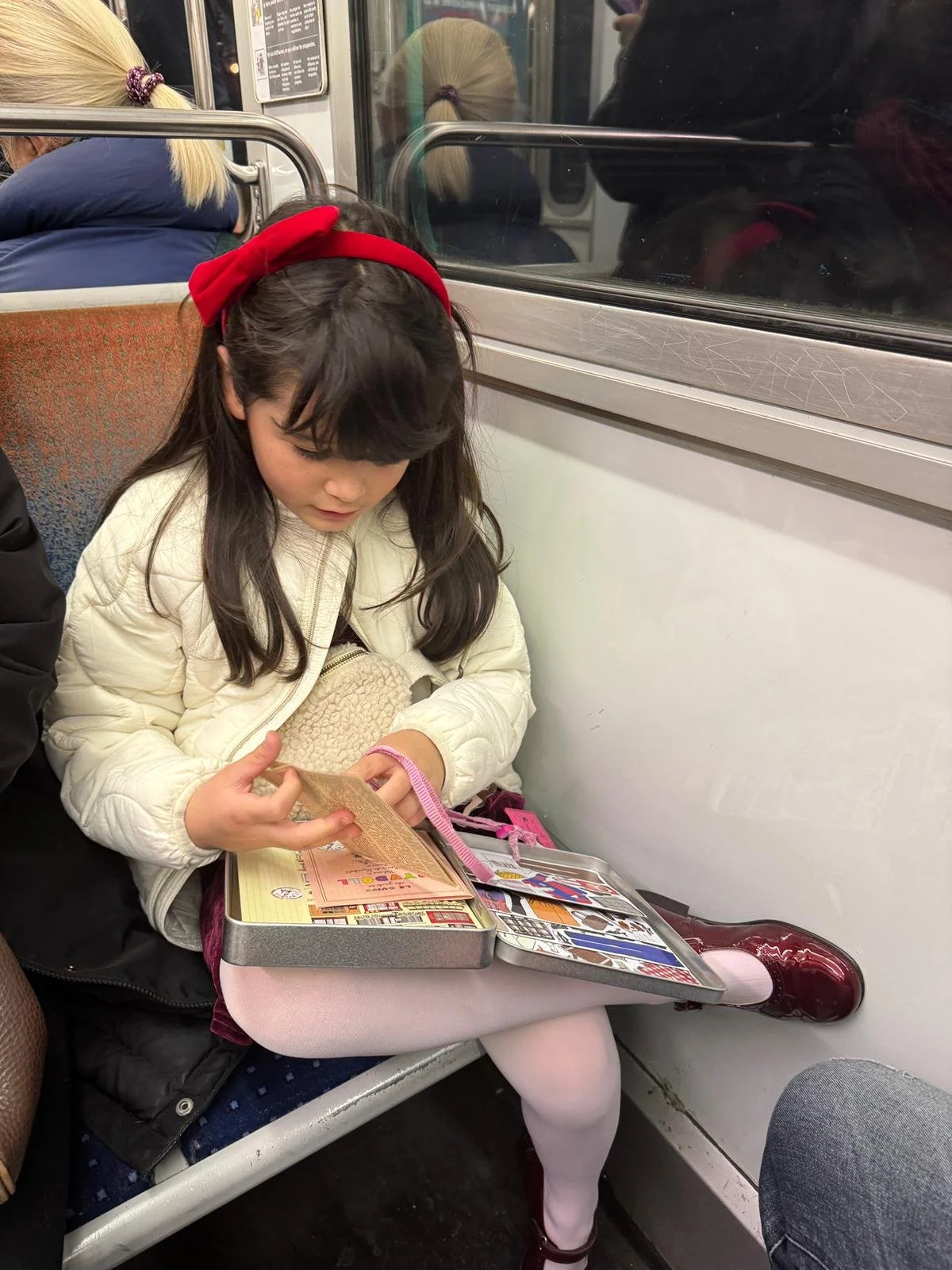 A young girl with a red headband sitting on a train seat, reading a book with a pink cover, while wearing a white jacket and pink tights, with a pink backpack on her lap.