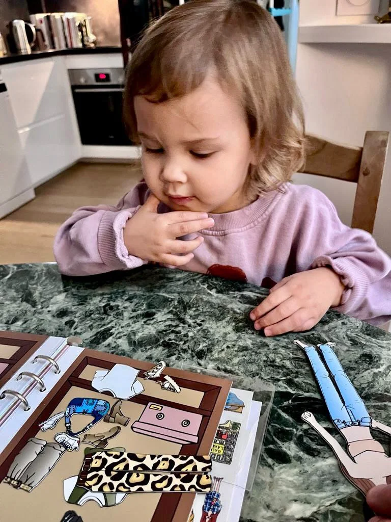 A young girl with brown hair in a pink sweatshirt sits at a green marble table looking at a sticker album with various paper cutouts of clothes and accessories.