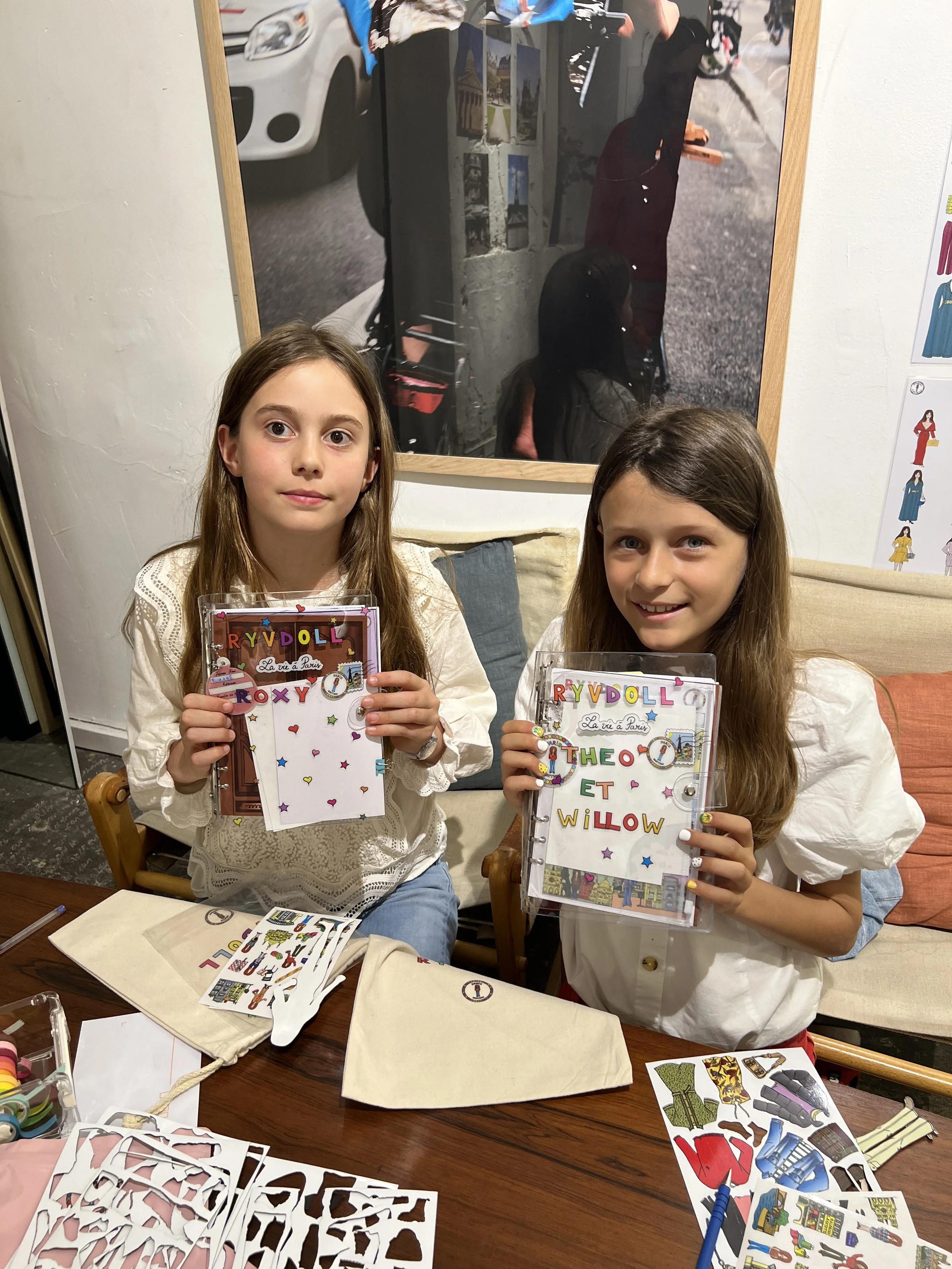 Two young girls sitting at a table holding colorful plastic folders with the names 'ROXY' and 'THEO ET WILLOW' on them. The table has craft supplies including stickers and cutouts, and the girls are smiling in a casual indoor setting.