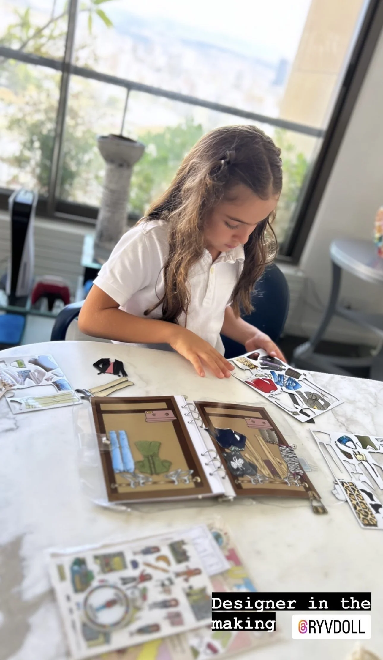 Young girl sitting at a table, looking at fashion design stickers and paper dolls with clothing options, in a room with large windows and a city view.