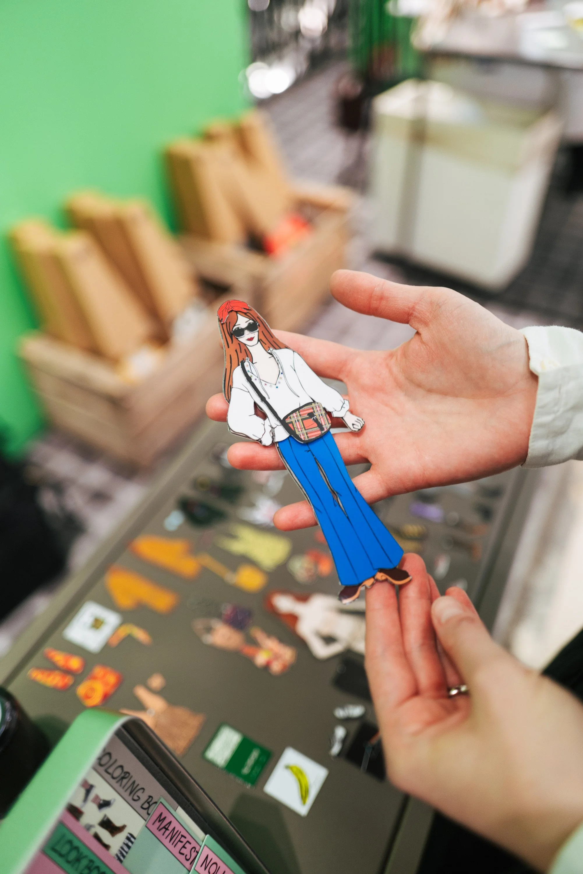 Person holding a colorful illustrated paper doll of a woman with red hair, sunglasses, white shirt, blue bell-bottom pants, and a checkered bag, in a store with various stickers and items on the counter in the background.