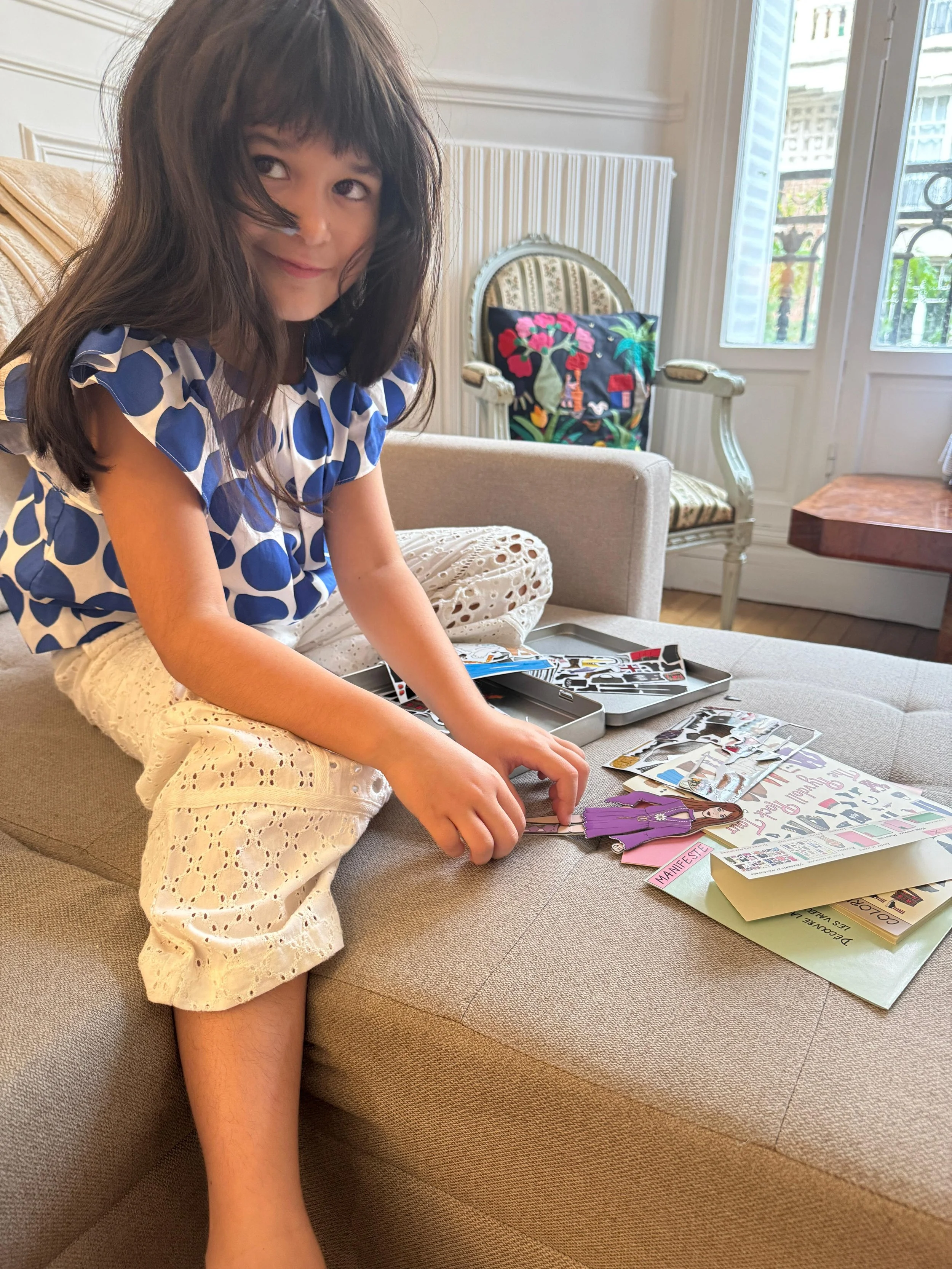 A young girl with long brown hair and a blue and white polka dot top, smiling while sitting on a beige couch, playing with paper cutouts and stickers, in a bright living room with large windows and outdoor greenery.