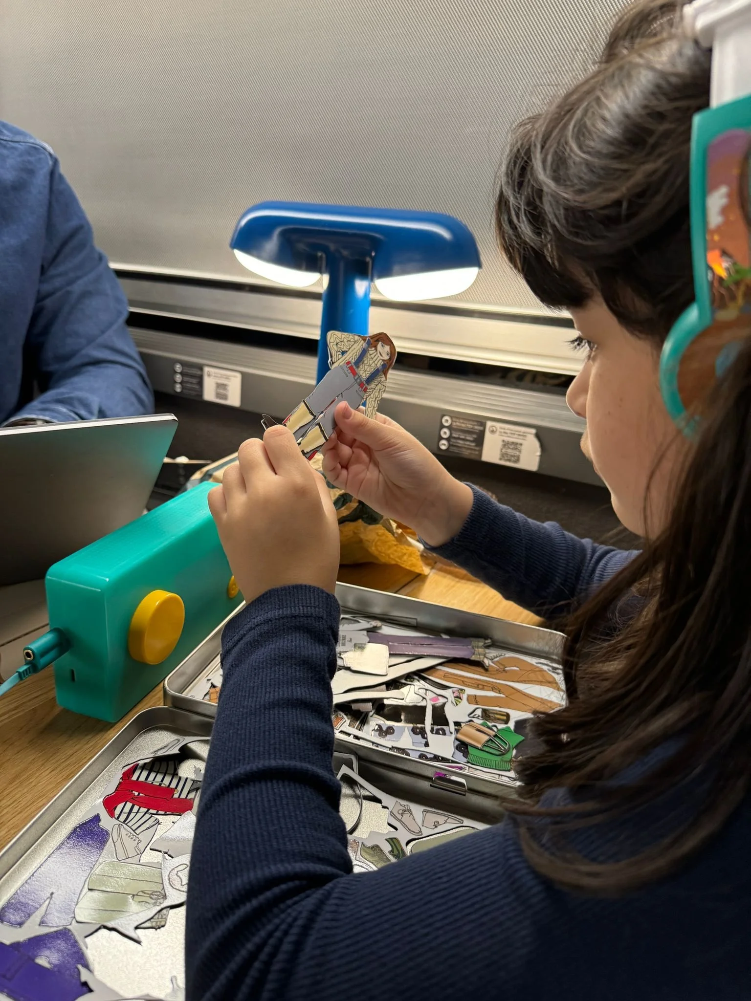 A girl with dark hair, wearing glasses and a navy blue long-sleeve shirt, is sitting at a table and holding a paper cutout of a person. She is surrounded by scissors, paper scraps, and other craft materials, with a study lamp shining overhead.