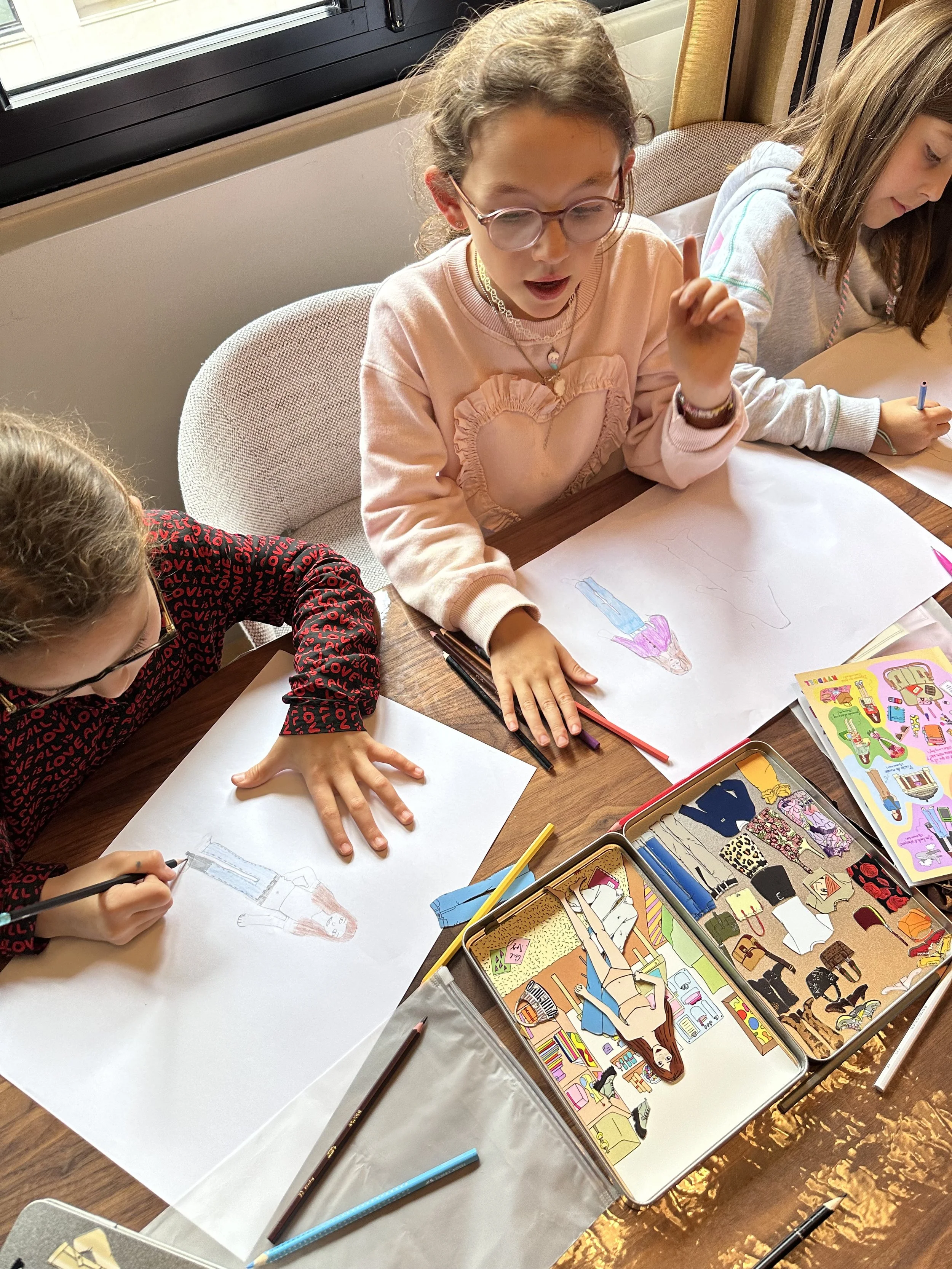Three young girls sitting at a table, drawing and coloring pictures of people on large sheets of paper, with coloring supplies and picture books nearby.