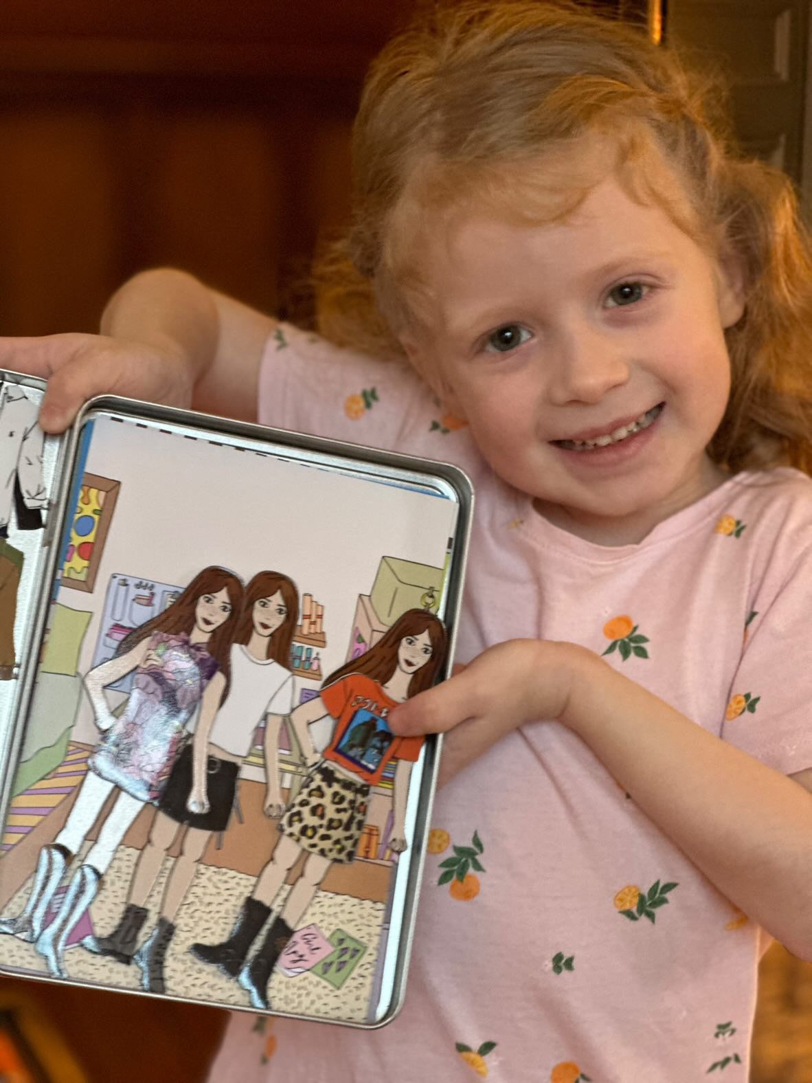 A young girl with red hair and a pink shirt with orange and green fruit prints, smiling and pointing at a cartoon image on her phone, which features three animated women in a room.