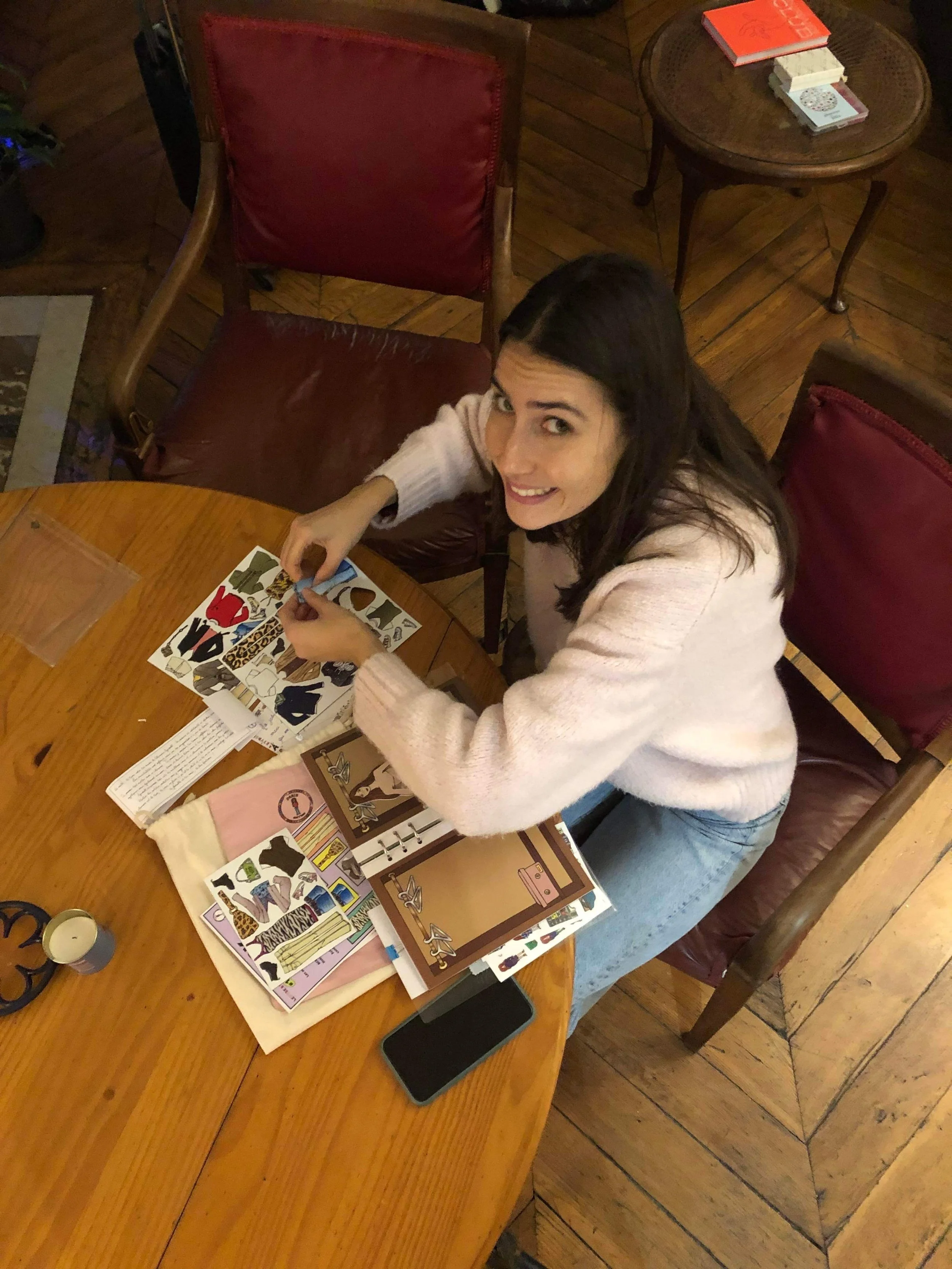 A woman with long dark hair sitting at a wooden table, looking up and smiling at the camera, working on a collage or scrapbook with colorful paper and stickers. The table has a black smartphone, a candle, and some papers on it. The setting appears to