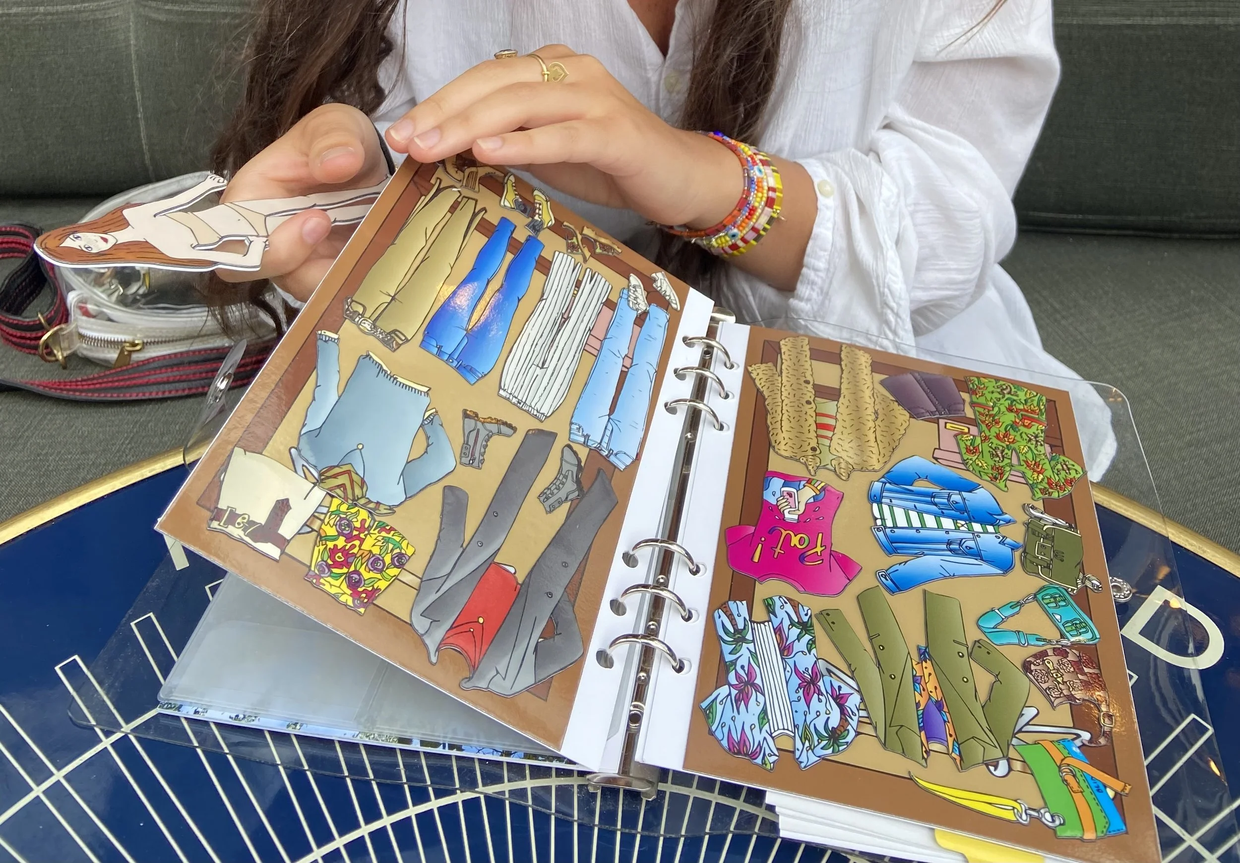 A person looking at a colorful illustrated closet with various clothing items and accessories in a photo album. The person is wearing bracelets and a gold ring, sitting on a sofa with a bag nearby.