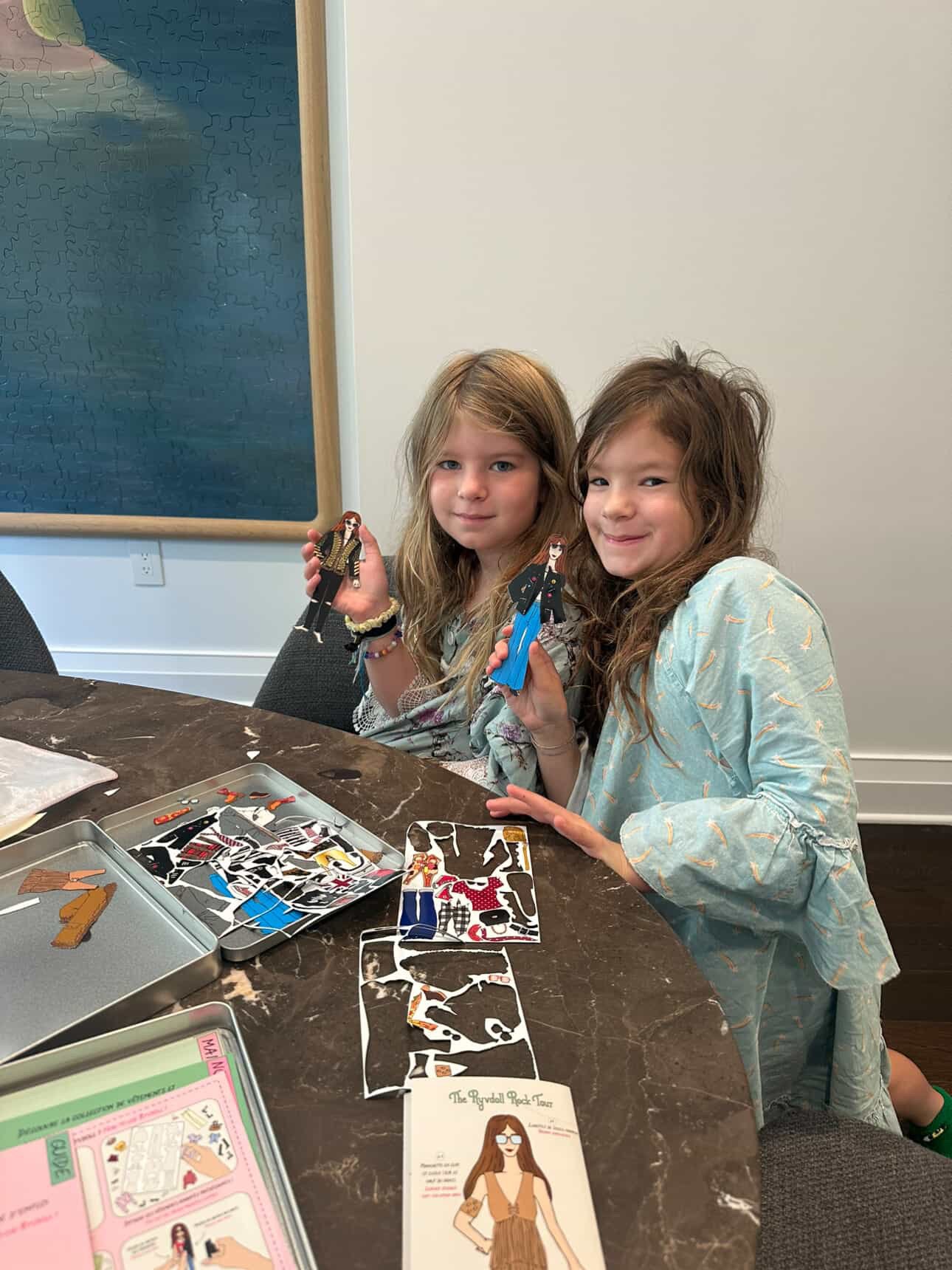 Two young girls sitting at a table, holding paper dolls, and working on a craft project with various cut-out paper pieces and instructions.