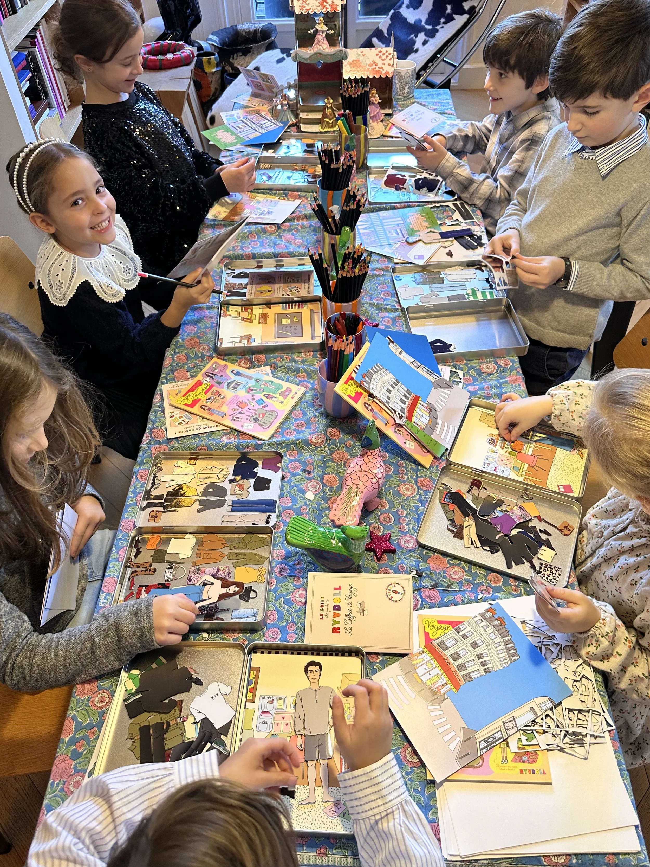 Children working on paper doll craft project at a table with colorful paper cutouts, art supplies, and comic books.