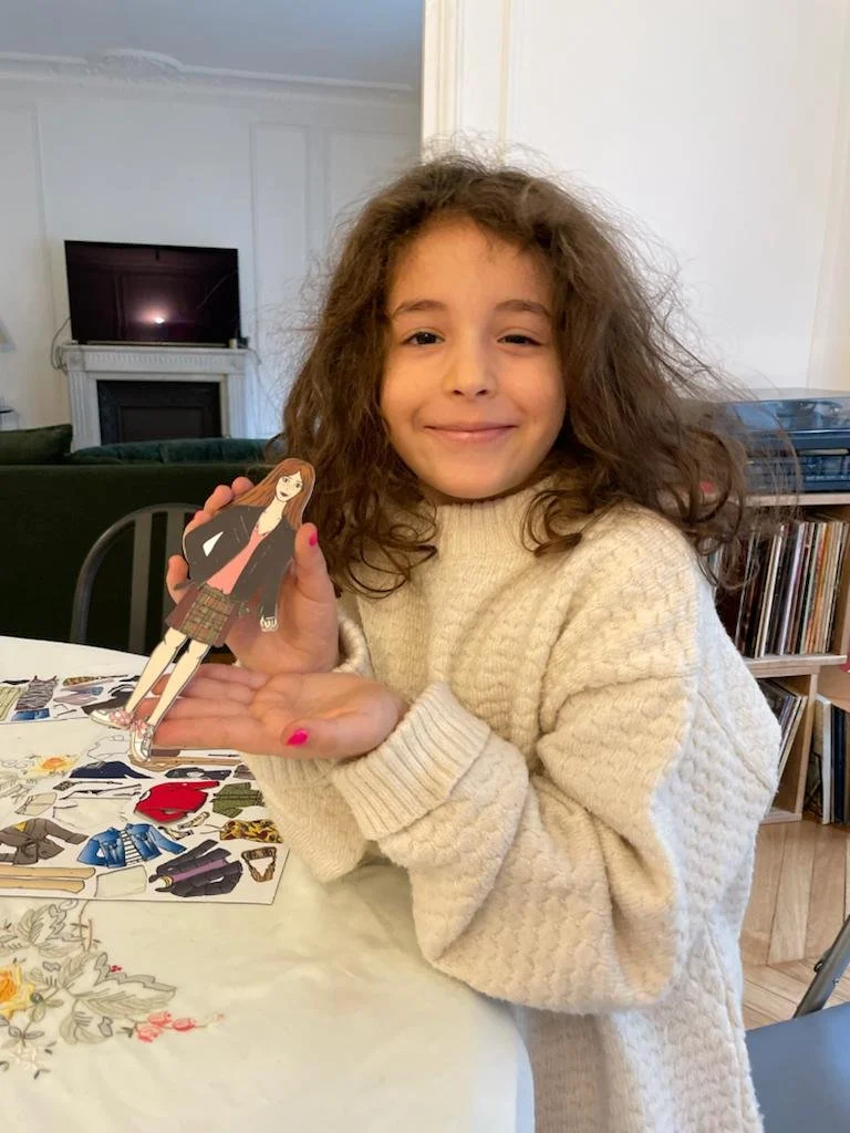A young girl with curly hair, wearing a beige sweater, smiling and holding a paper doll of a girl with long hair, wearing a black blazer and a plaid skirt, sitting at a table with cut-out clothing and accessories.