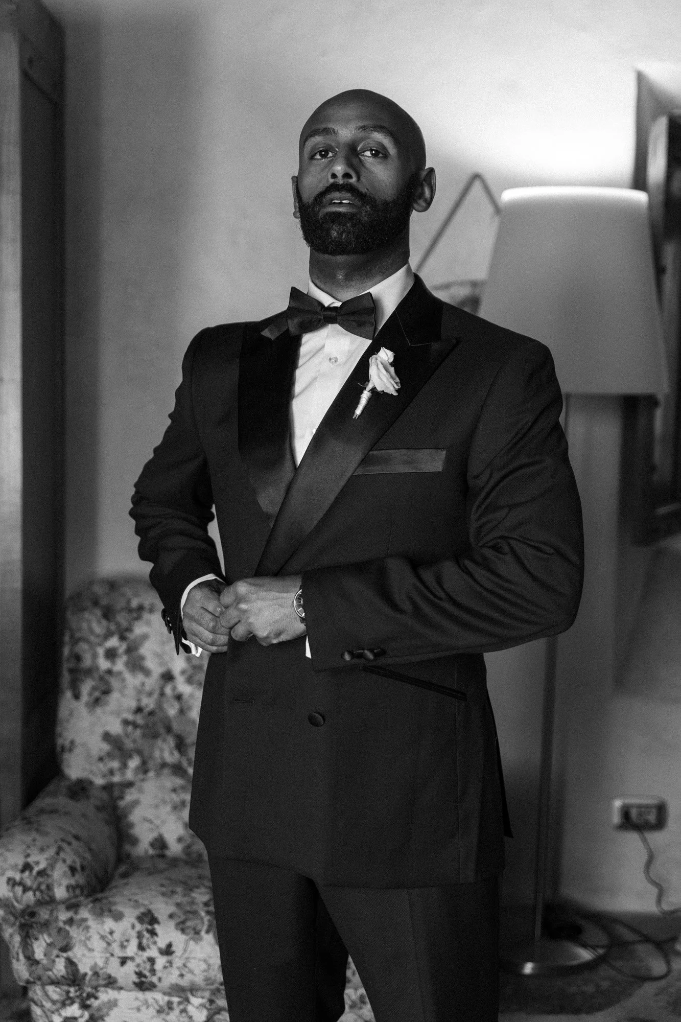 Uomo elegante con smoking nero, farfalla e fiore sulla giacca, posa in modo formale in una stanza condivisa.