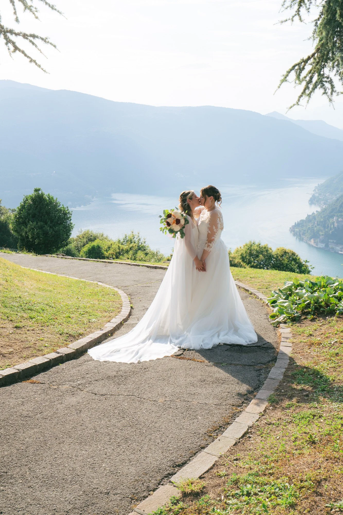Due donne in abiti da sposa si baciano davanti a un paesaggio lacustre montano con alberi e vegetazione, sotto un cielo luminoso.