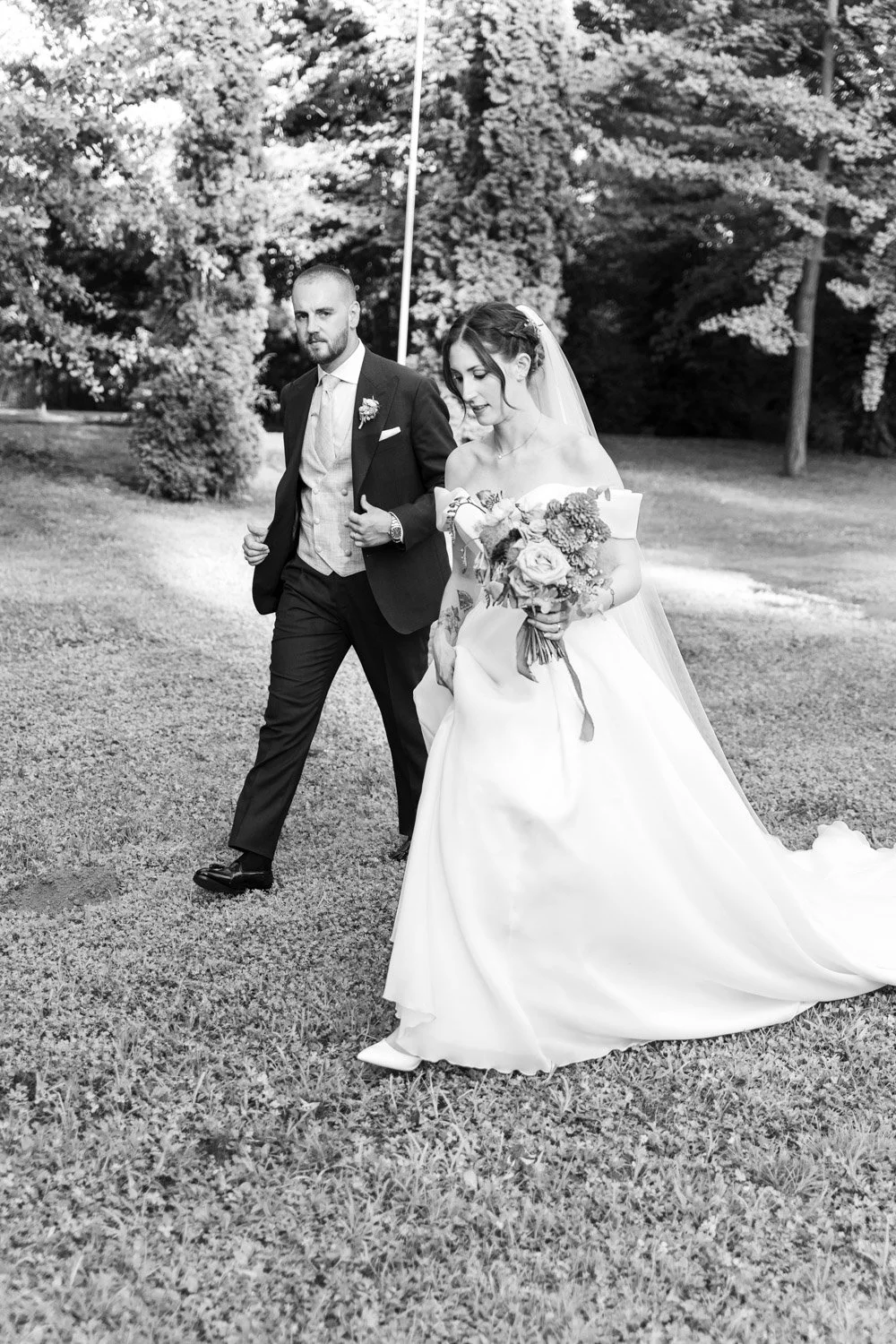 Una coppia di sposi cammina all'aperto, con lo sfondo di alberi e natura, l'uomo indossa un abito da cerimonia scuro e la donna indossa un abito da sposa bianco con un bouquet di fiori.