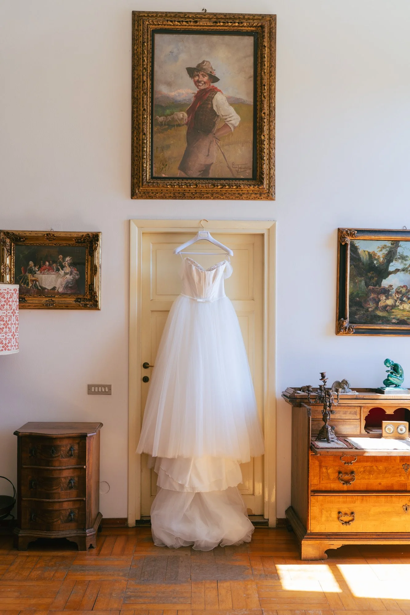 Una stanza con pareti chiaro e quadri appesi, un vestito da sposa bianco appeso su una porta, mobili in legno antico e un quadro sopra la porta raffigurante un bambino sorridente in abiti rustici.