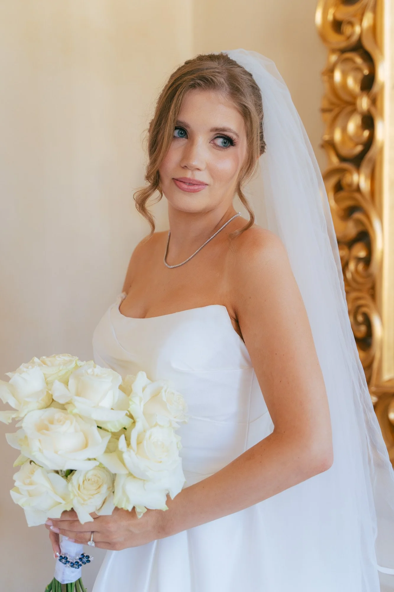 Una sposa con abito bianco, con un bouquet di rose bianche, guarda verso sinistra, con uno sfondo neutro e un dettaglio dorato a destra.