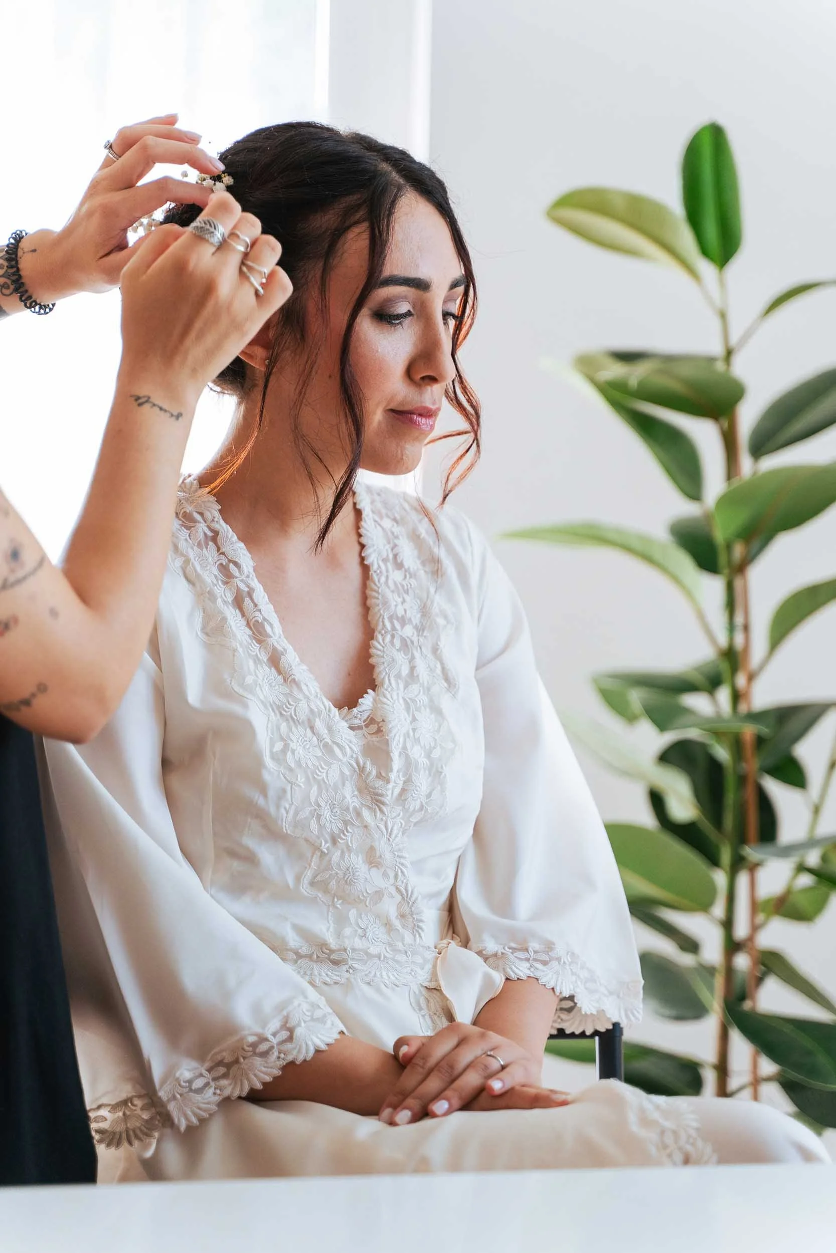 Una donna con capelli scuri e ondulati è seduta con le mani in grembo, mentre una persona sta sistemando i suoi capelli. La donna indossa un vestito bianco con dettagli in pizzo.